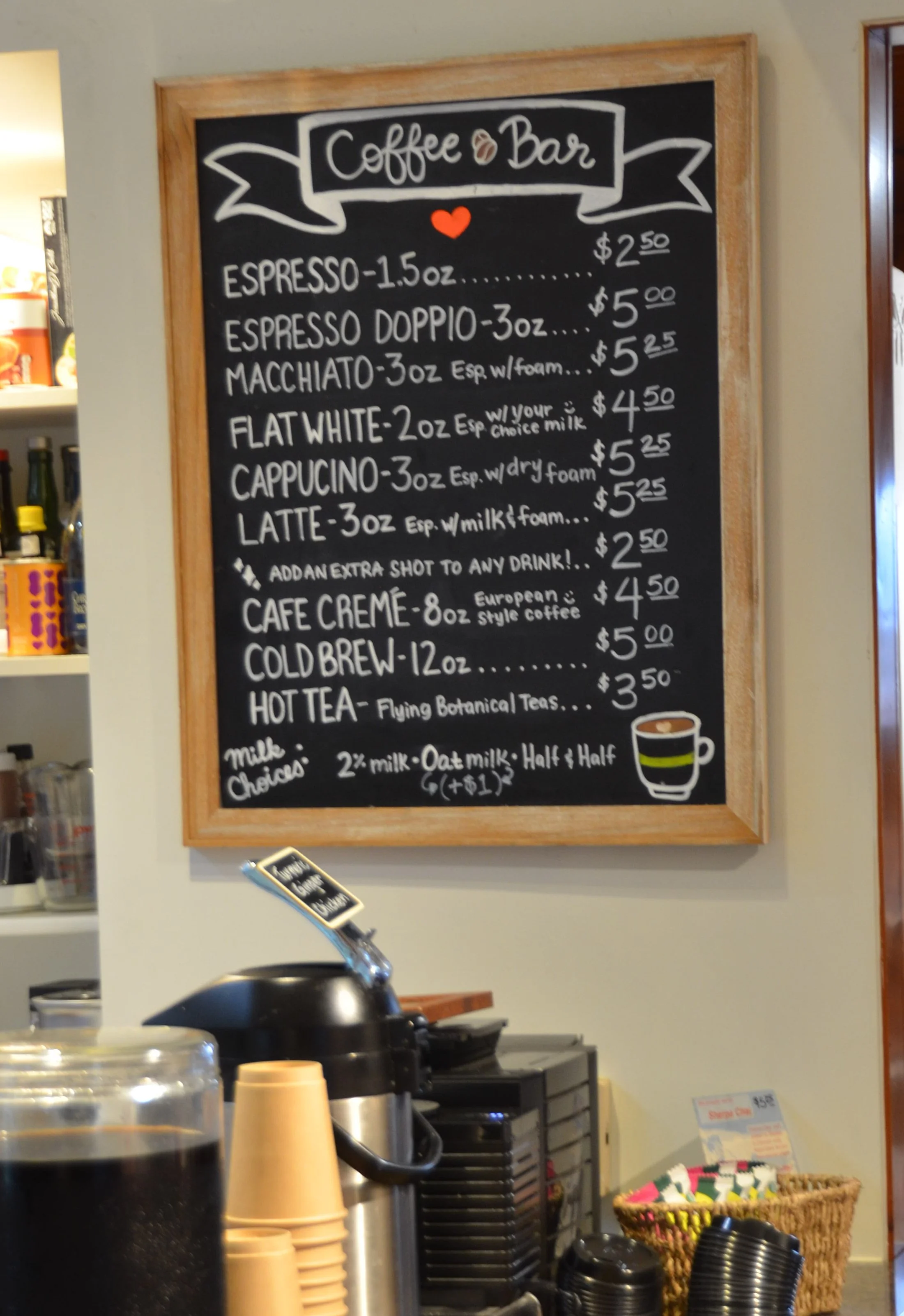 A black chalkboard menu with a wooden frame lists coffee drinks and their prices at a coffee shop. Items include espresso, doppio, macchiato, flat white, cappuccino, latte, café crème, cold brew, hot tea, and various milk options.