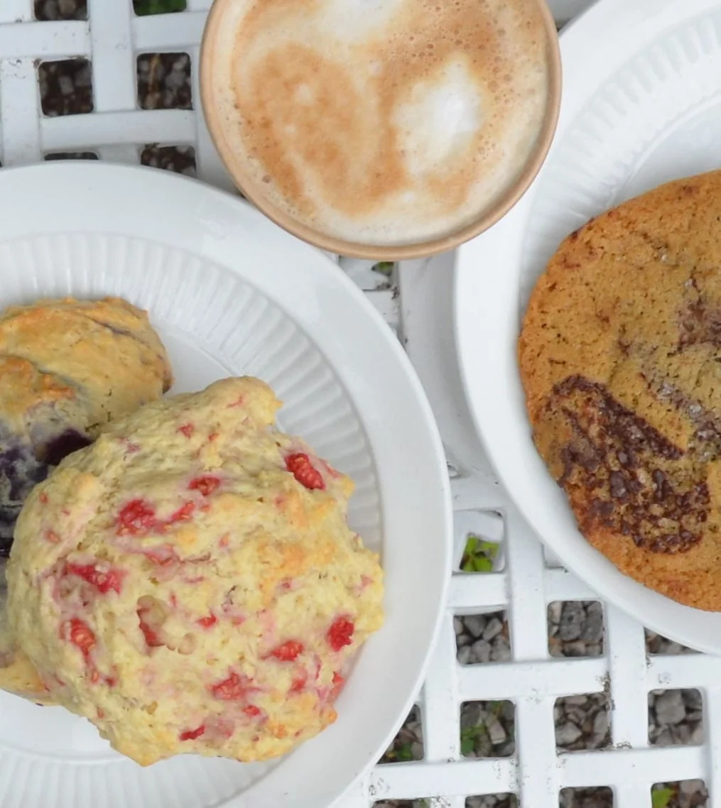 Scones with berries on a white plate, chocolate chip cookie on a white plate, and a cup of frothy cappuccino on a white table.