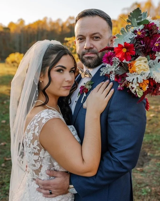 A bride and groom on their wedding day standing outdoors. The bride is wearing a lace wedding dress and a veil, while the groom is in a blue suit with a bow tie. They are holding a large bouquet of colorful flowers. The background features trees and 