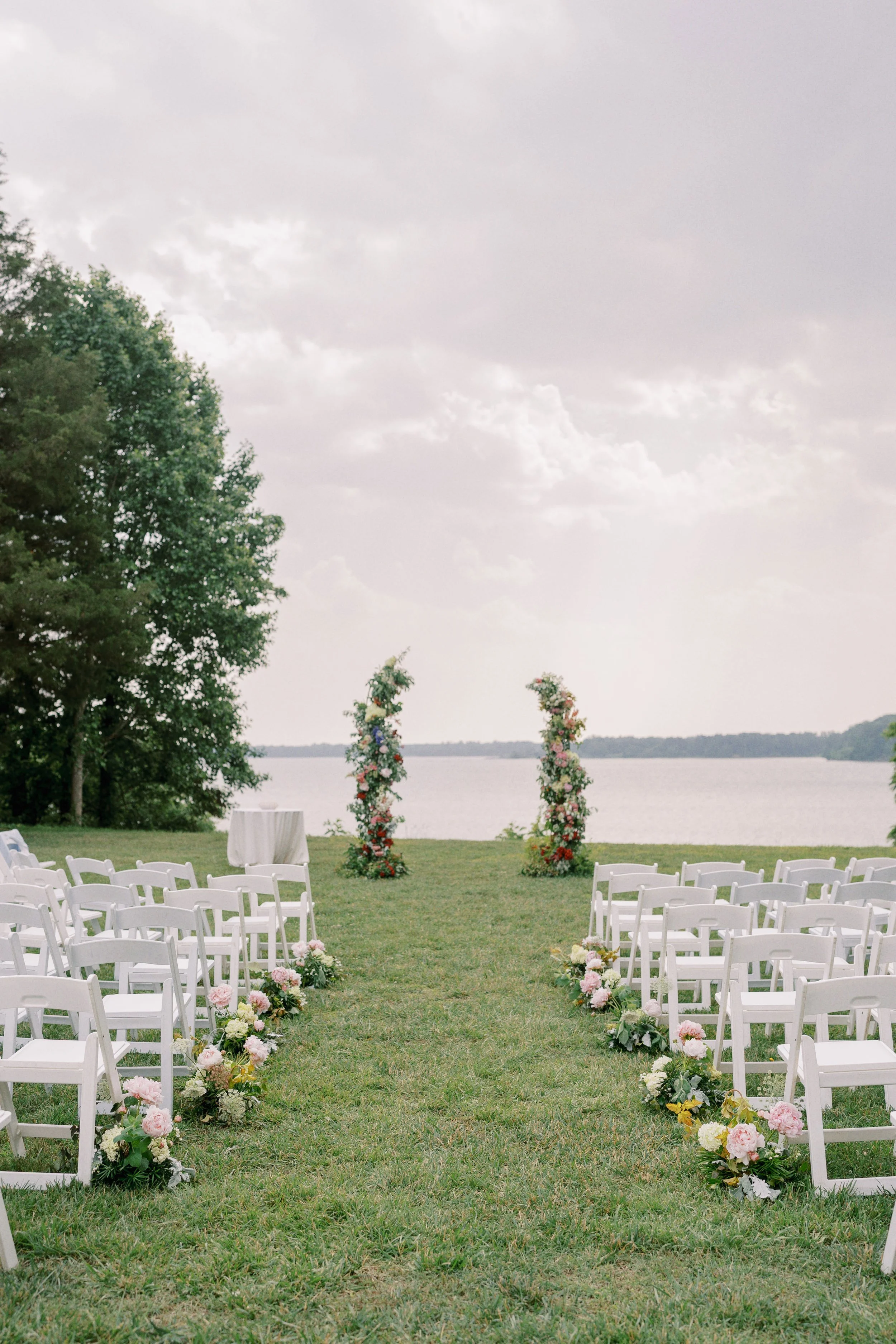 Outdoor wedding ceremony setup on a grassy area near a body of water, with white chairs arranged in rows on either side of an aisle decorated with flowers, and a floral arch at the end of the aisle.