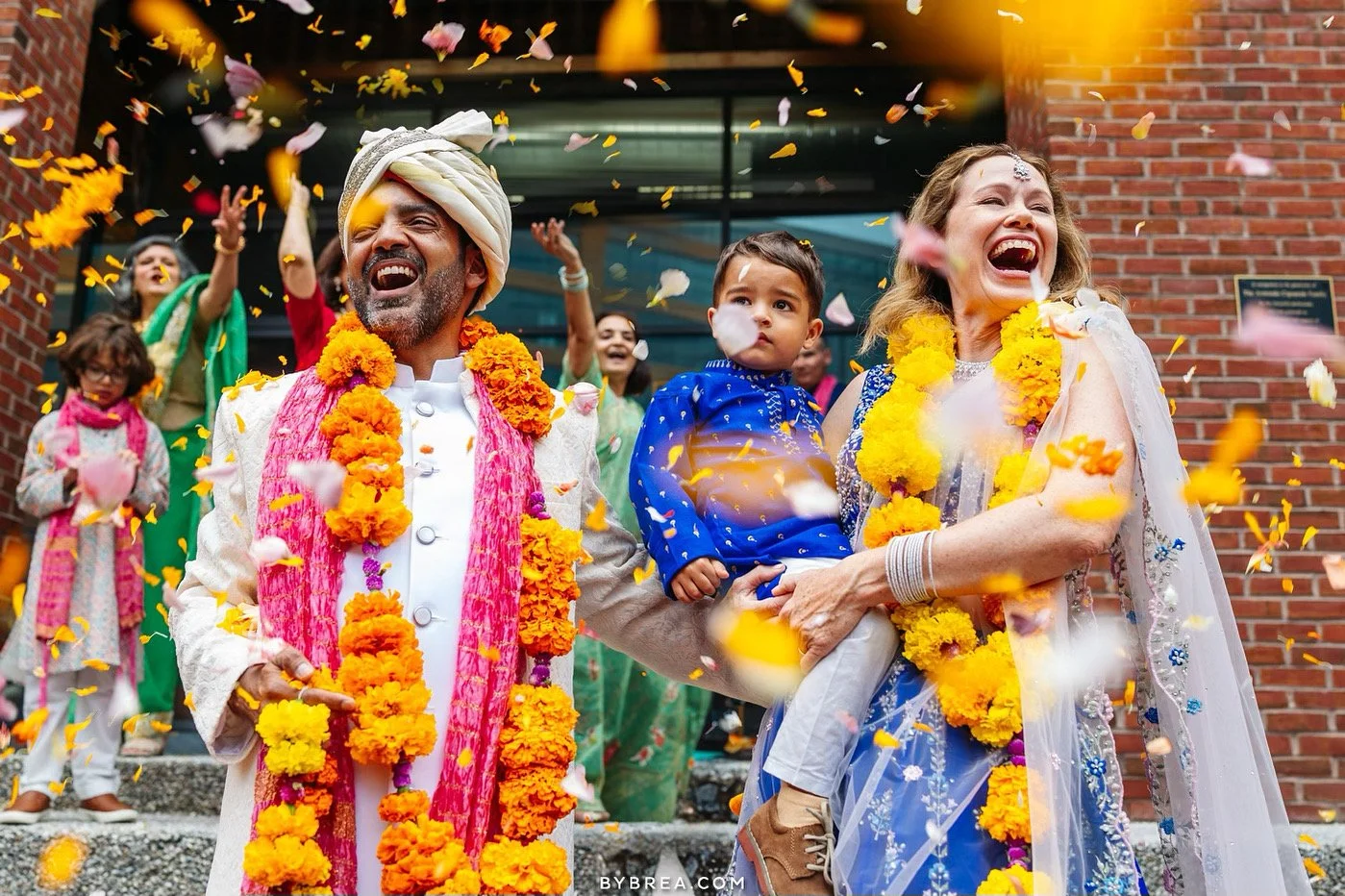 A celebration scene featuring a joyful group of people dressed in traditional Indian attire, with marigold flower garlands, as flower petals are thrown in the air outside a building with a brick wall.