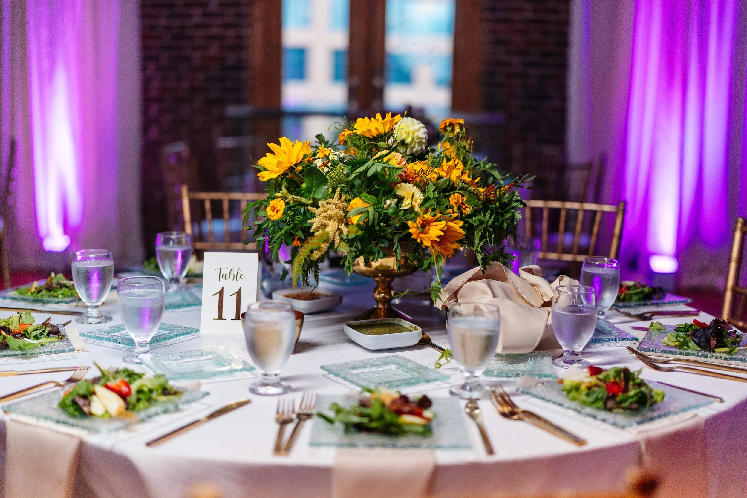 A wedding or formal event table with a large floral centerpiece of sunflowers, white and yellow flowers, and greenery. The table is set with salad plates, glasses of water, gold utensils, and a table number sign indicating Table 11. Purple uplighting decorates the background with brick walls and curtains.