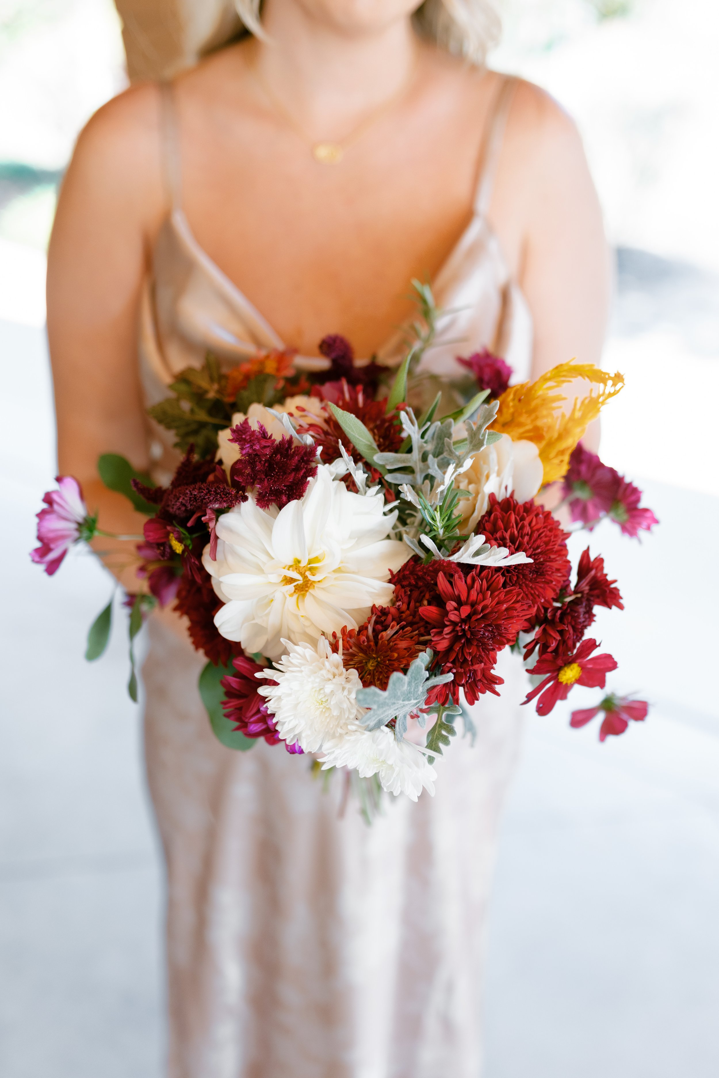 Person in a beige dress holding a colorful bouquet of white, red, pink, and yellow flowers.