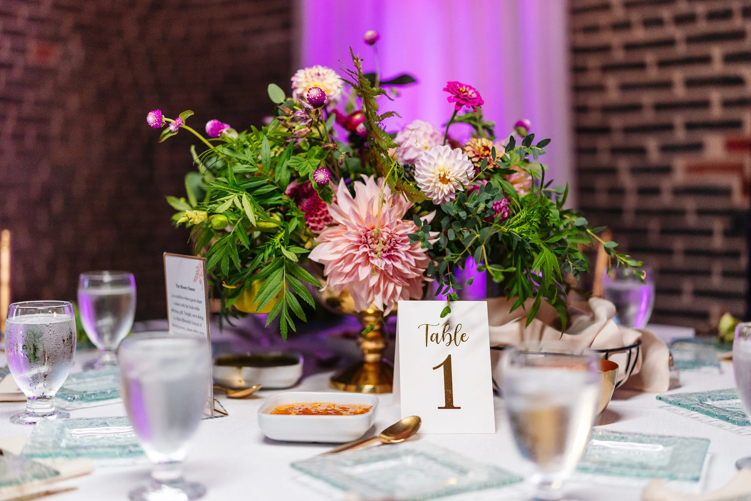 Elegant table setup with a floral centerpiece, water glasses, a bowl of soup, and a table sign labeled 'Table 1'. Wall with purple lighting in the background.