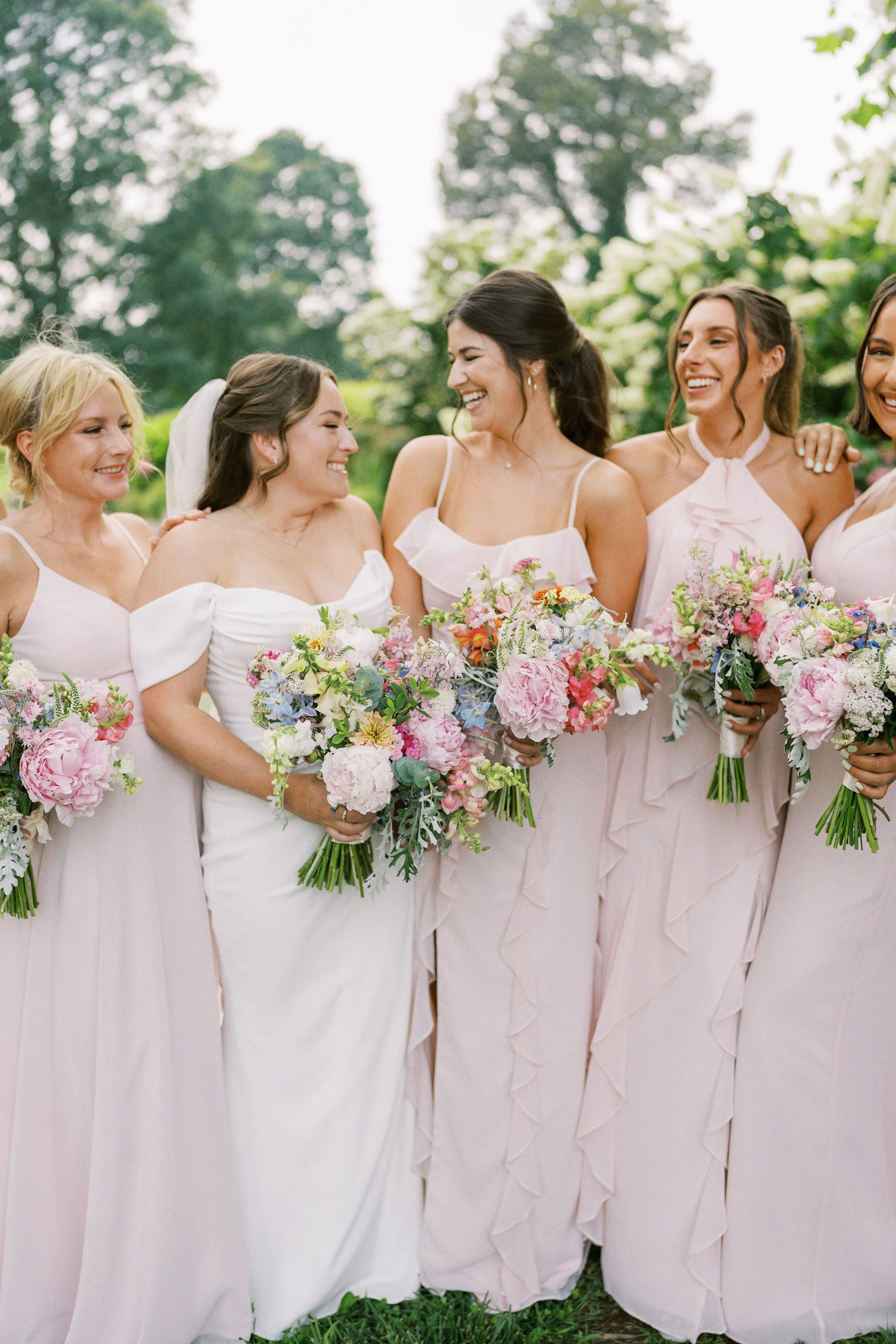 Group of women in pastel pink dresses holding flower bouquets, standing outdoors with trees in the background, smiling and interacting with each other.
