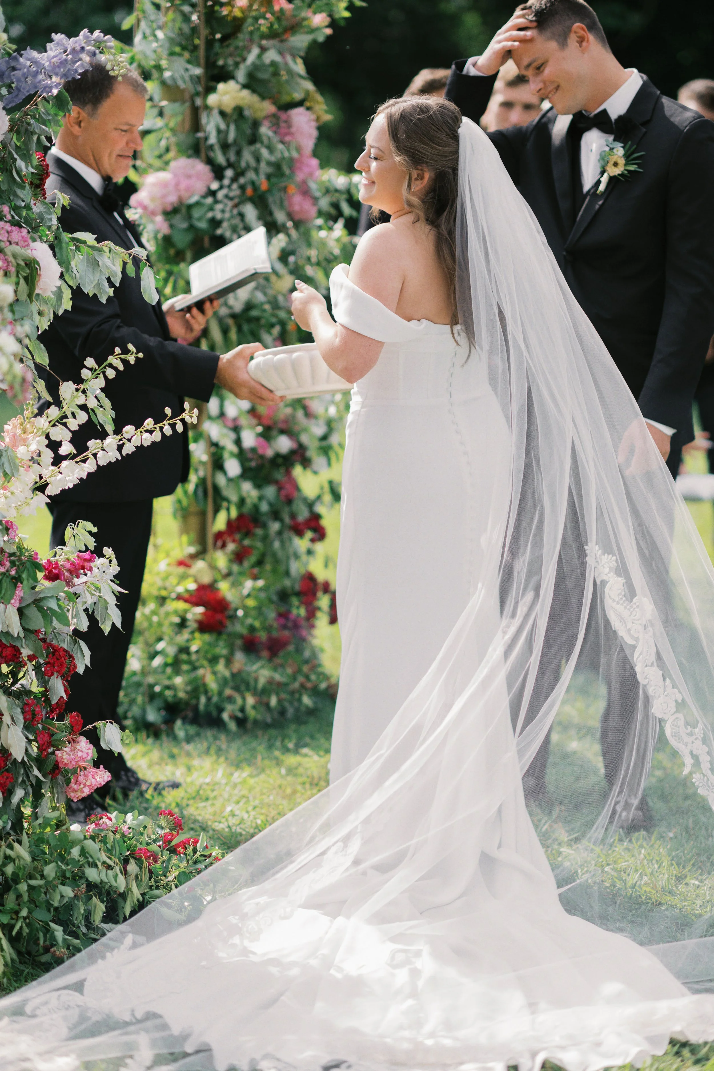 Bride and groom exchange vows during outdoor wedding ceremony with officiant, surrounded by flowers.