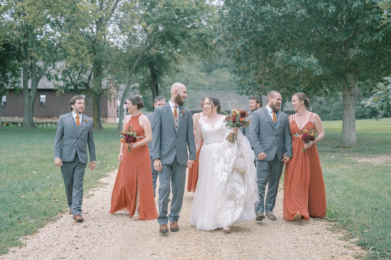 A group of eight people dressed in wedding attire walking outdoors on a dirt path, surrounded by green trees and grass. The bride is holding a bouquet and wearing a white lace wedding dress, while the bridesmaids hold bouquets and wear long rust-colo