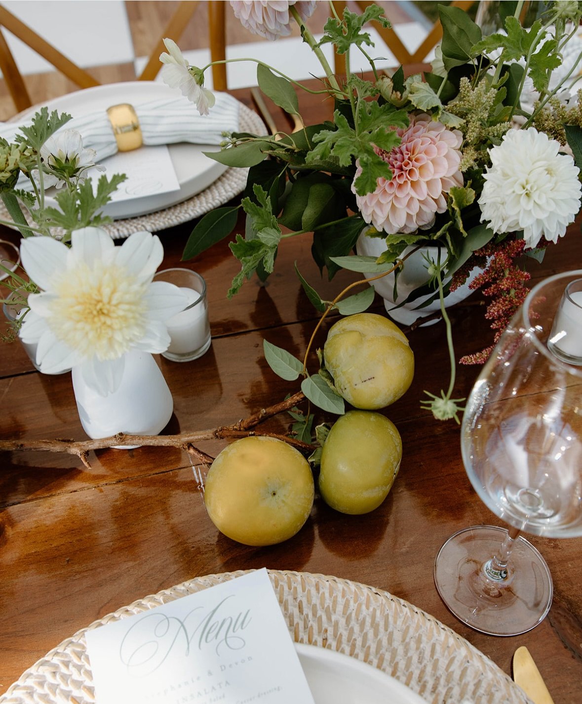 A dining table decorated with a floral centerpiece, apples on a branch, a menu, and place settings with candles.