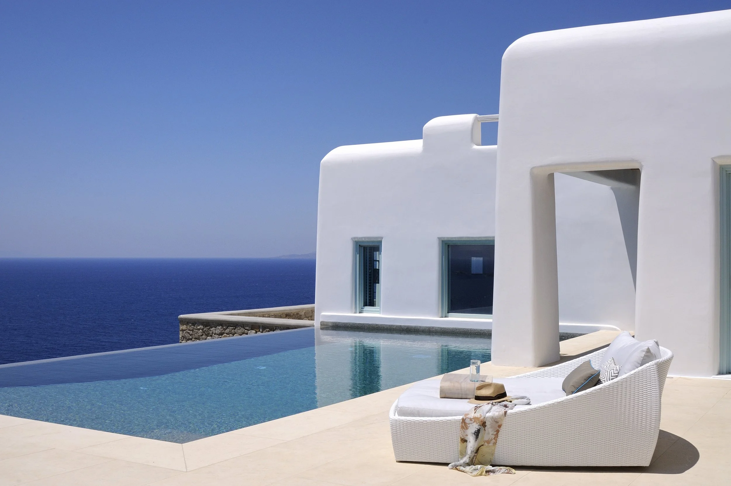White modern house with curved architecture and blue windows next to an infinity pool overlooking the ocean under a clear blue sky, with a white sun lounger with pillows, hat, and towel on beige tile flooring.