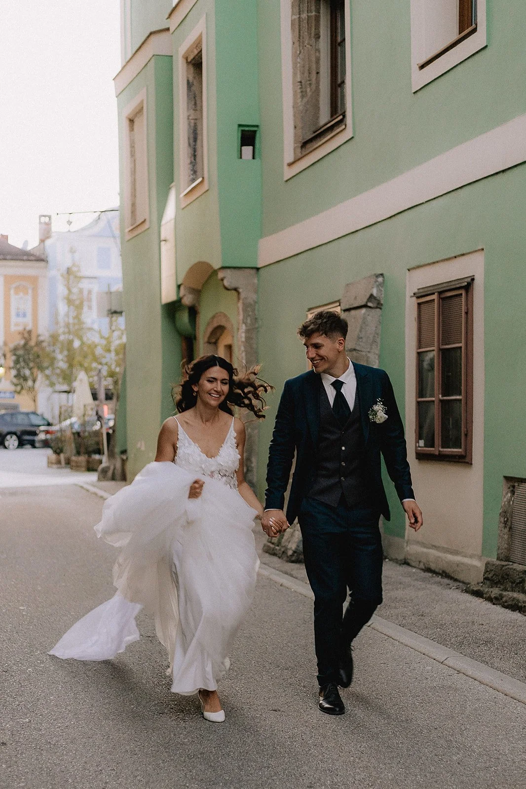 A bride and groom are walking hand in hand on a city street, smiling and enjoying their wedding day. The bride is wearing a white wedding dress, and the groom is dressed in a dark suit with a boutonnière.