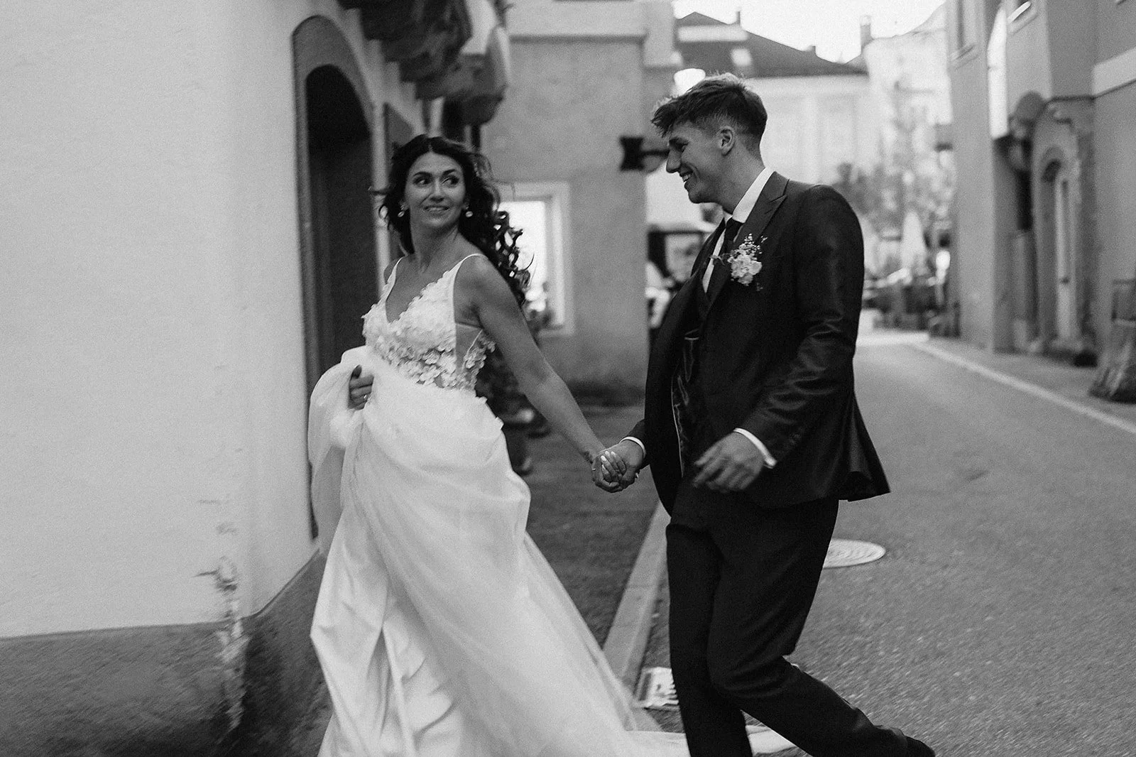 A bride and groom holding hands and smiling on a city street, with the bride in a wedding dress and the groom in a dark suit.