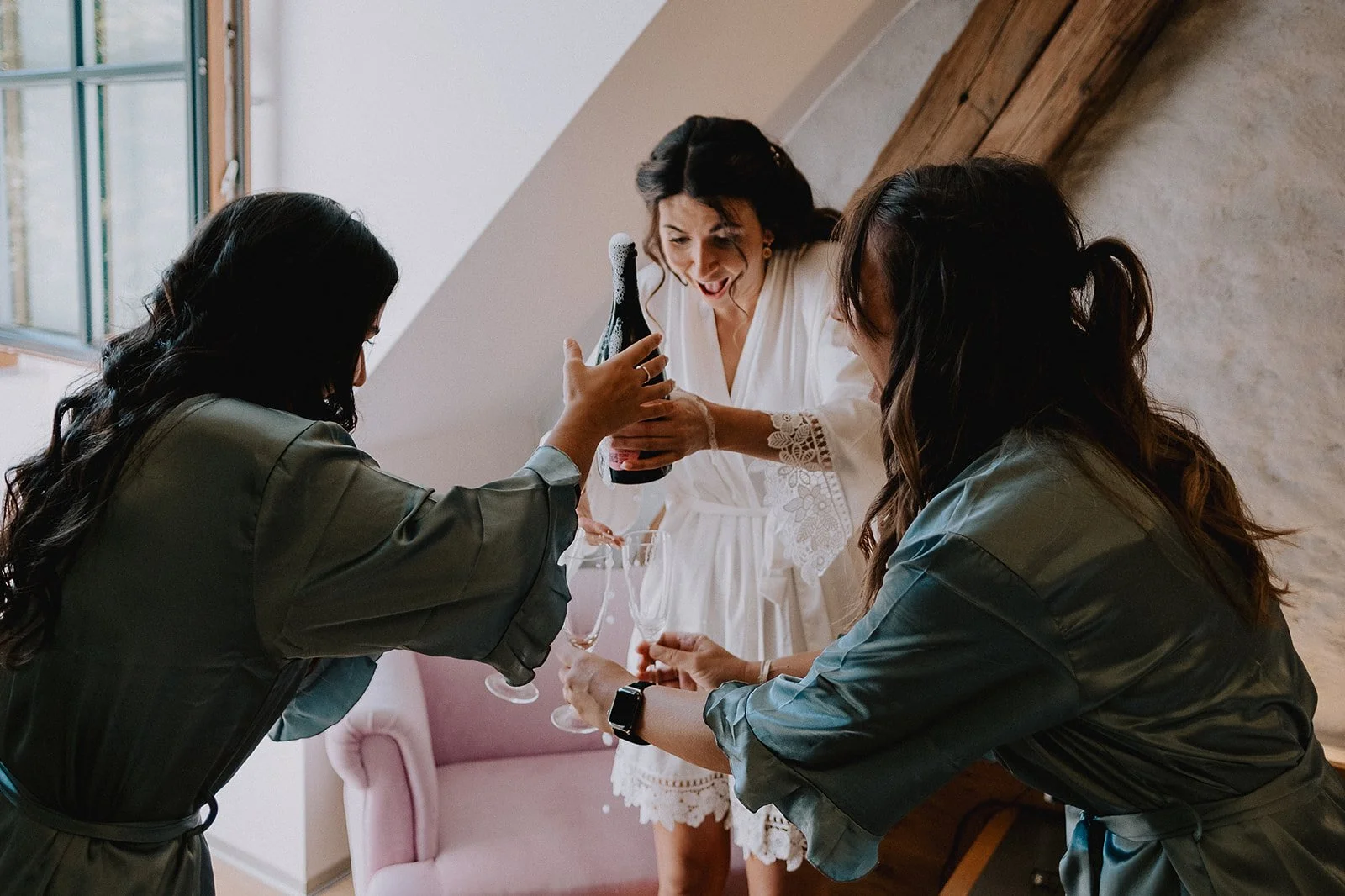 Three women celebrate, one woman in a white dress, two women in matching robes, toasting with champagne bottles and glasses in a cozy room with a pink chair and wooden beams.