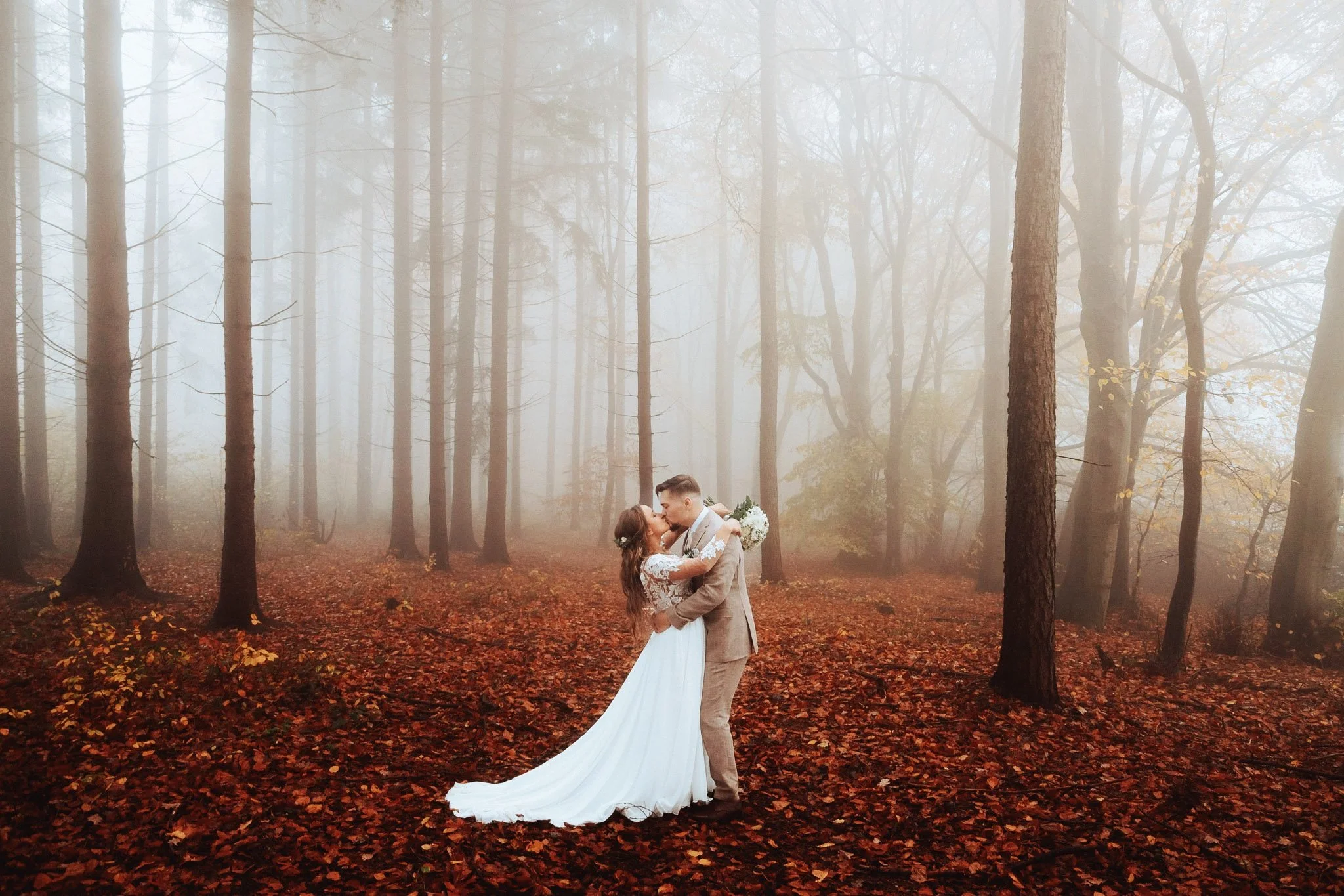 A bride and groom kissing in a foggy forest, with fallen autumn leaves covering the ground
