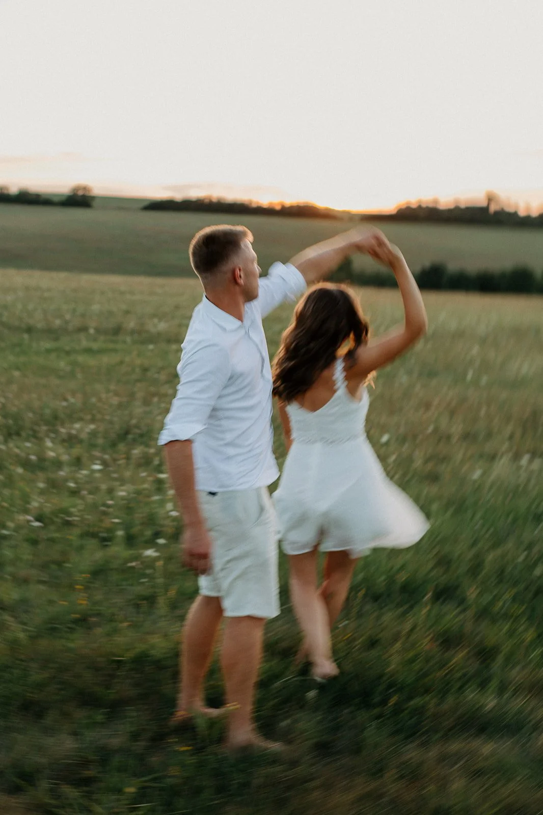 A couple dancing in a grassy field during sunset, with the man in a white shirt and shorts, and the woman in a white dress.