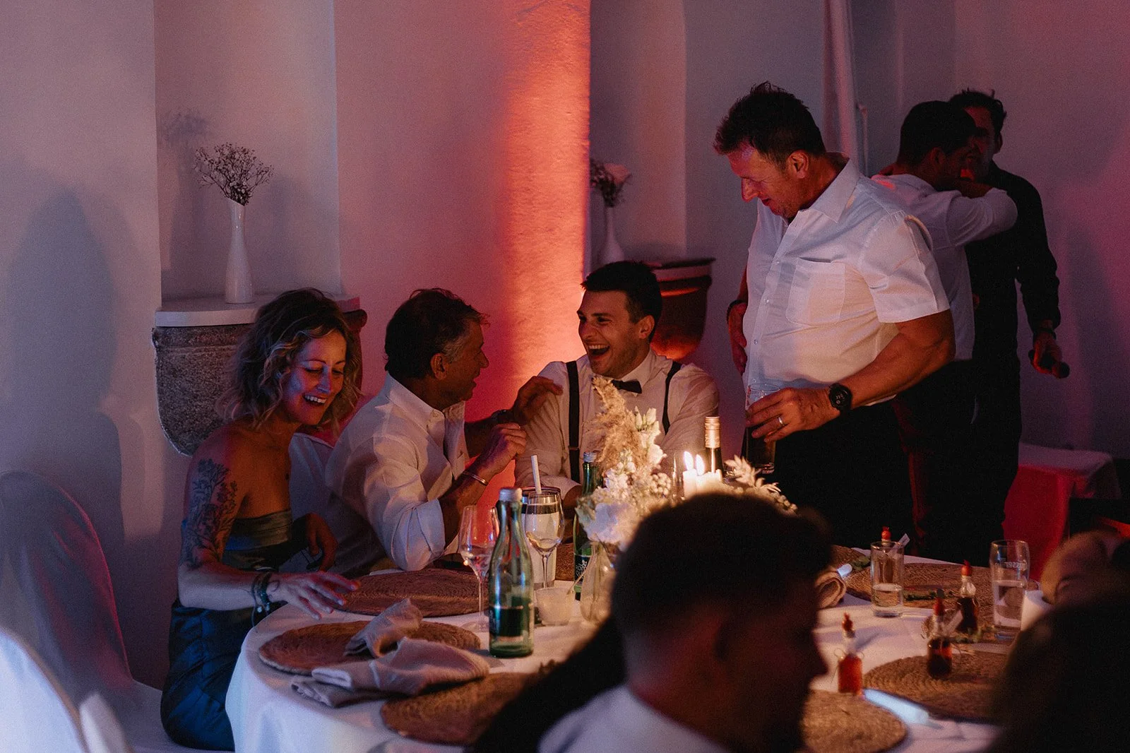 People celebrating at a dinner party, laughing and smiling around a table with candles and drinks in a dimly lit room with warm lighting.