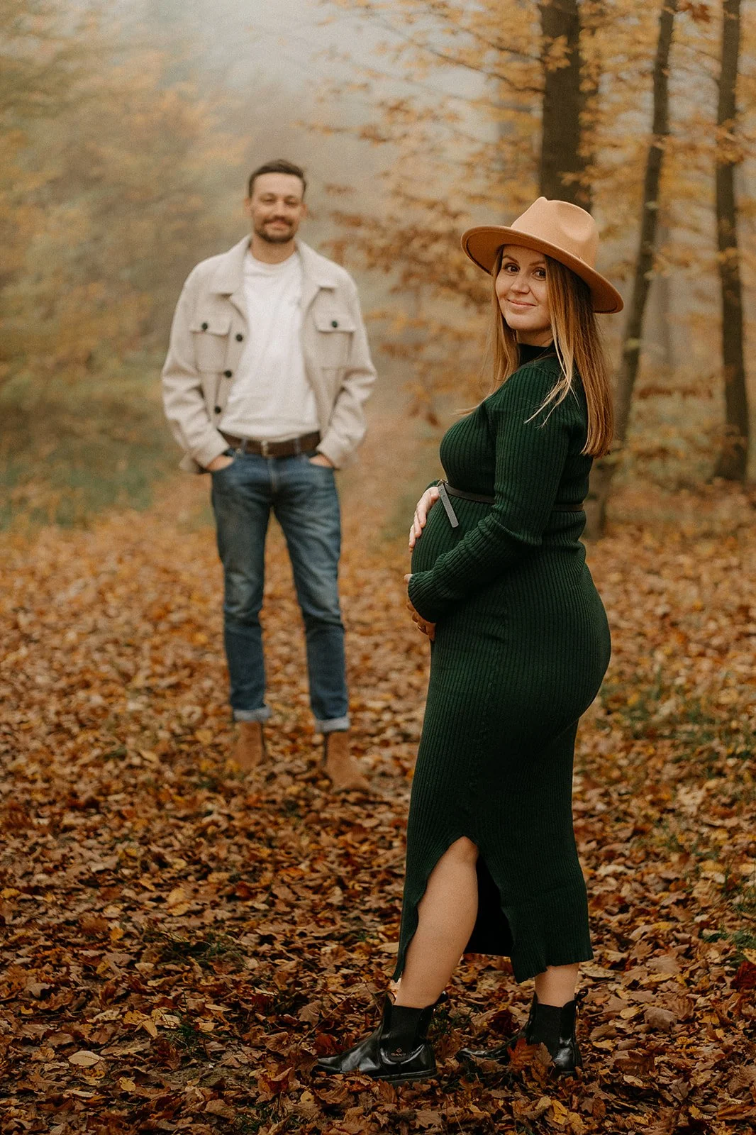 A pregnant woman wearing a green dress, black boots, and a tan hat is standing in the foreground, smiling and looking at the camera, with a man in a white jacket and blue jeans standing in the background in an autumnal forest.