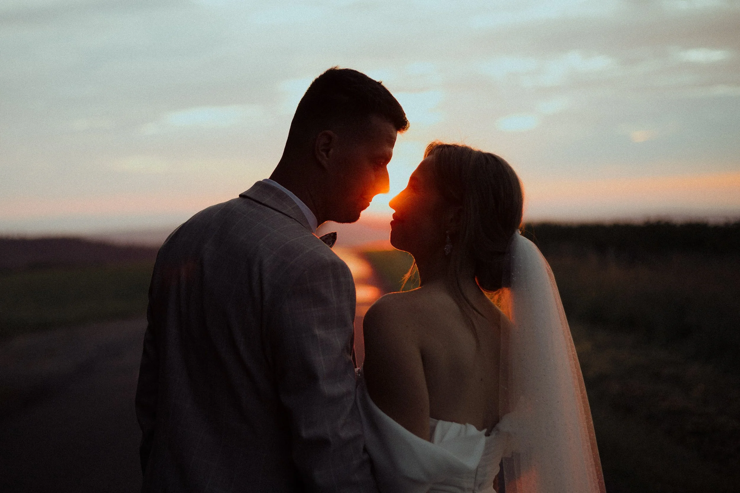 A bride and groom standing close together at sunset, with the sun between their faces, on a rural path with fields in the background.