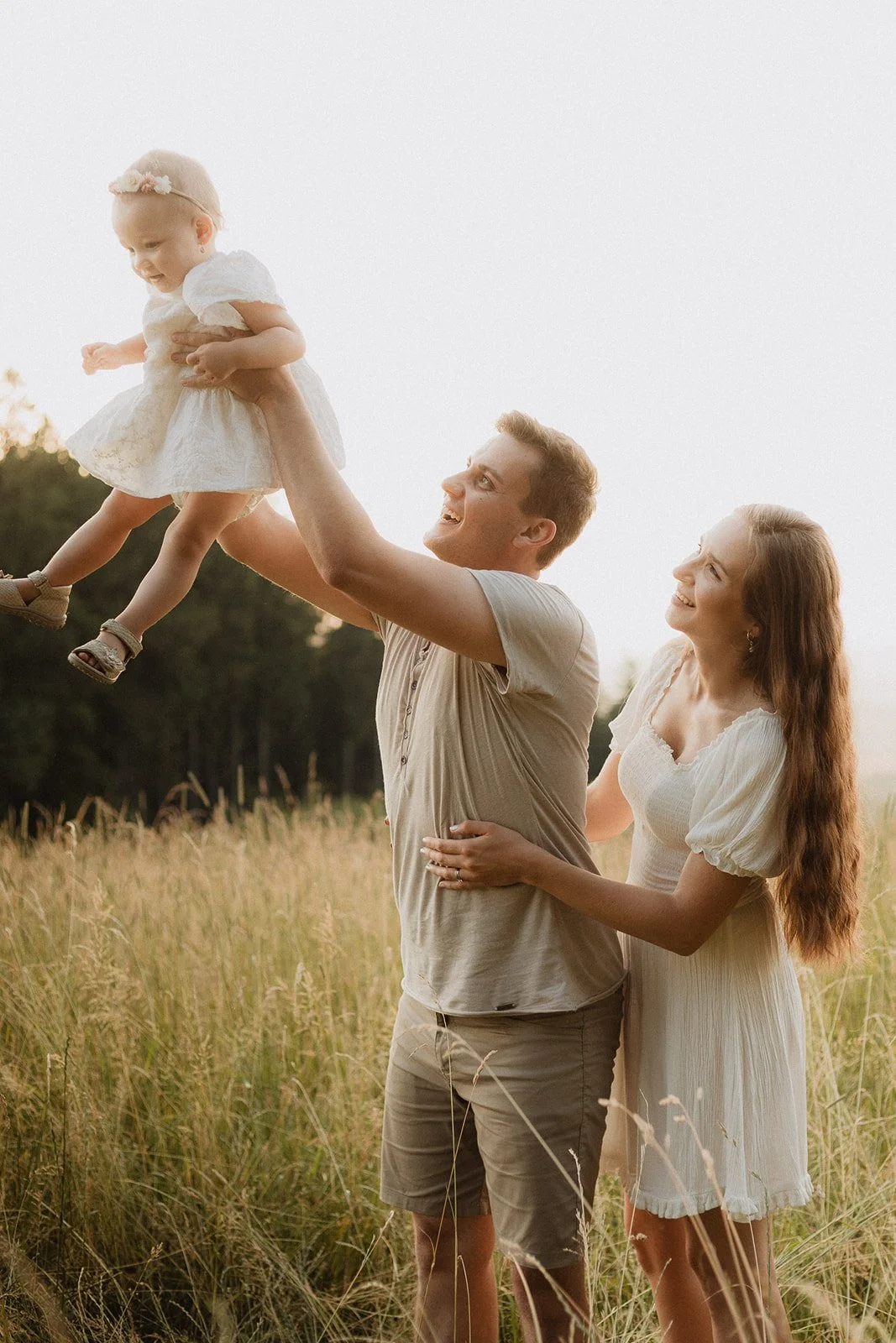 A young family enjoying a moment outdoors in a grassy field during sunset, with the father lifting a toddler girl wearing a white dress and a headband, while the mother watches and smiles.