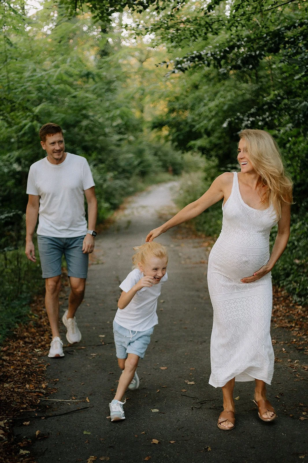 A family of three, including a woman, a man, and a young girl, are walking along a wooded trail. The woman is wearing a white dress and sandals, the man is in a white t-shirt and shorts, and the girl is in a white t-shirt and shorts. They are smiling and appear to be enjoying a playful moment together.
