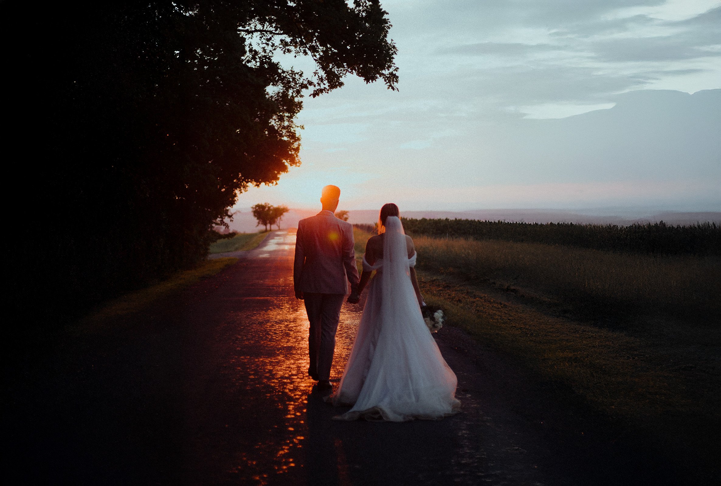 A bride and groom walking hand in hand on a rural road at sunset, with the bride wearing a white wedding dress and veil, and the groom in a suit, holding a bouquet of flowers.