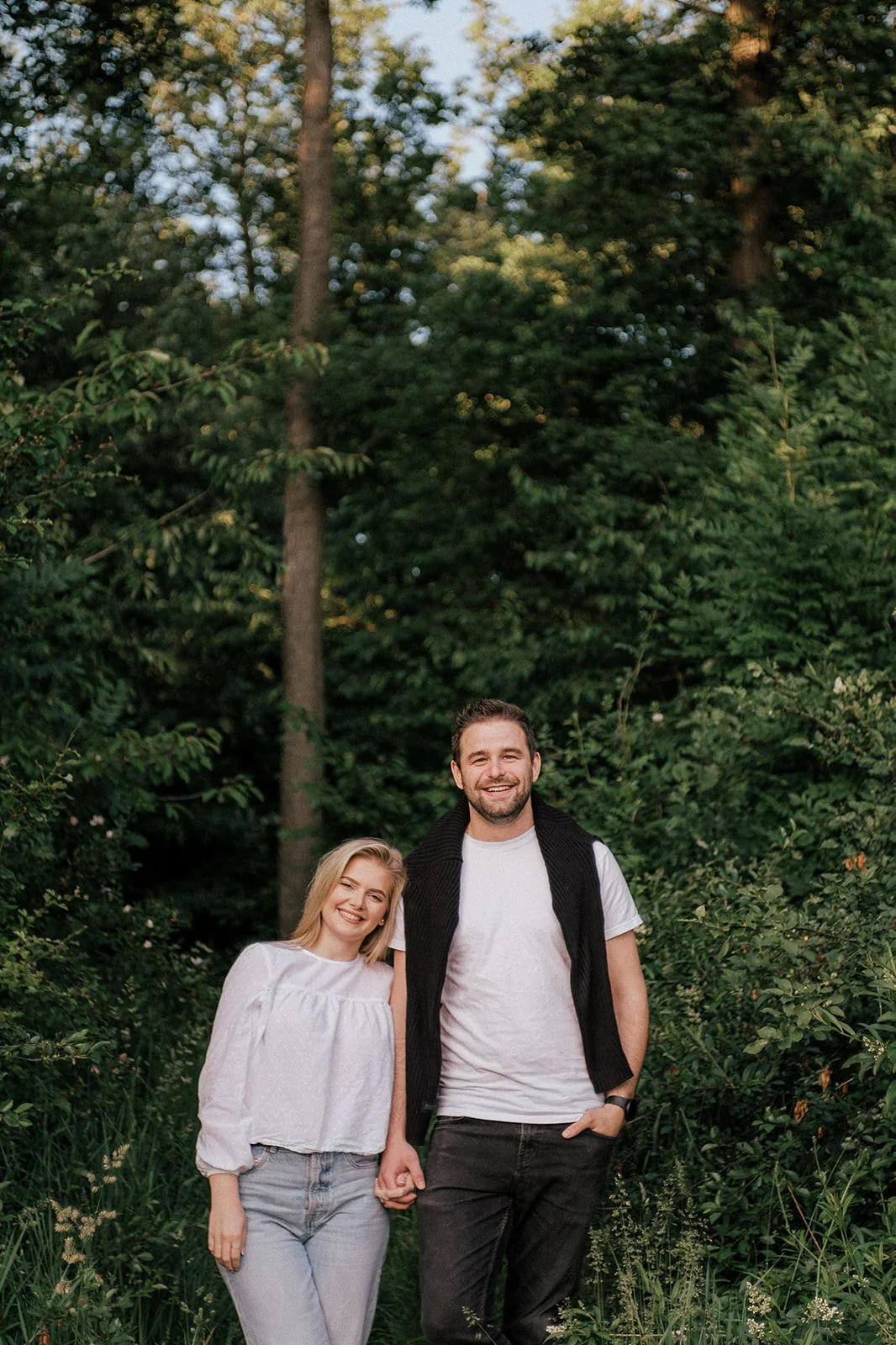 A smiling couple walking hand in hand through a lush green forest.