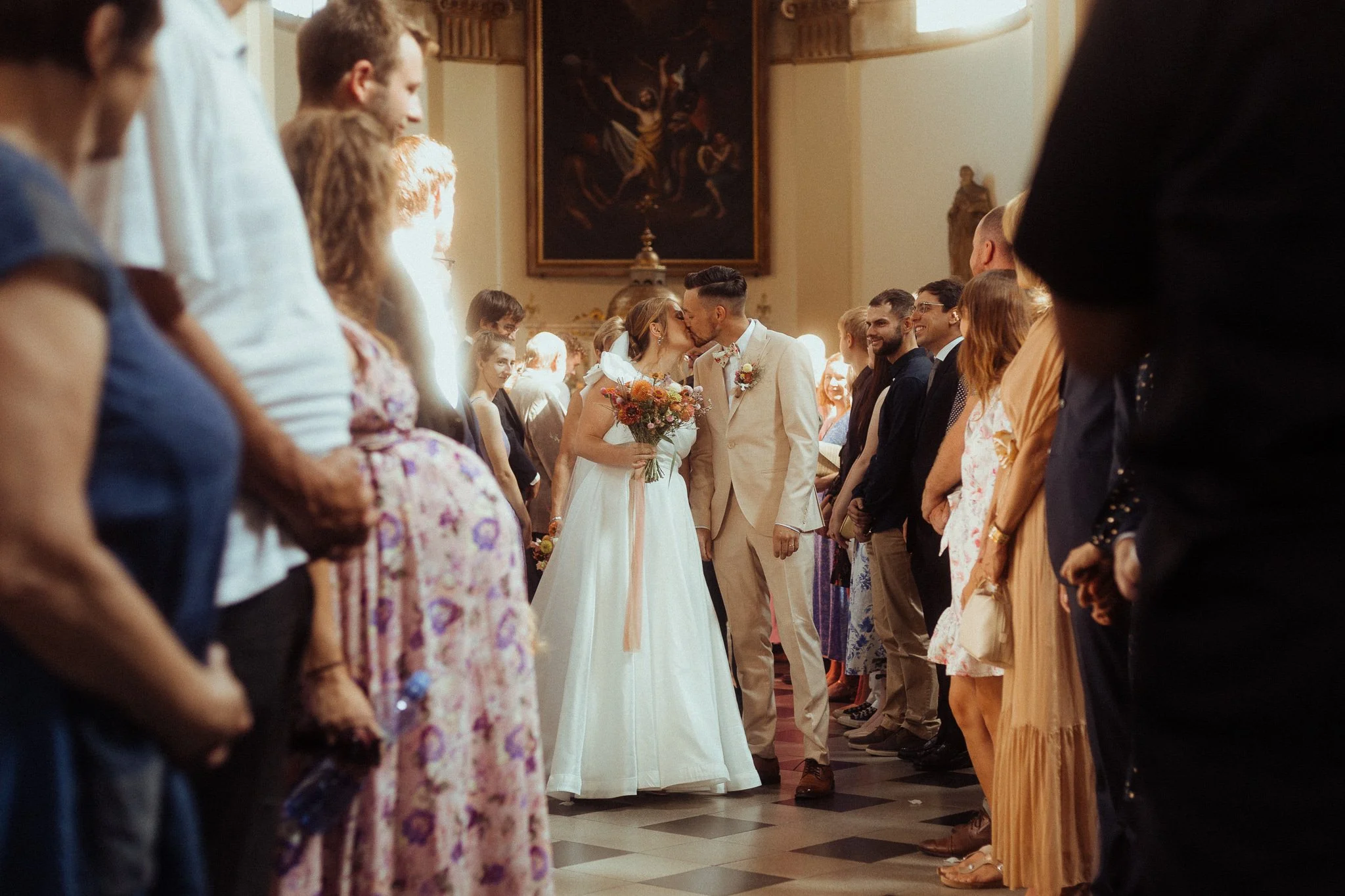 A newlywed couple is kissing in a church while surrounded by wedding guests standing in a row on either side.