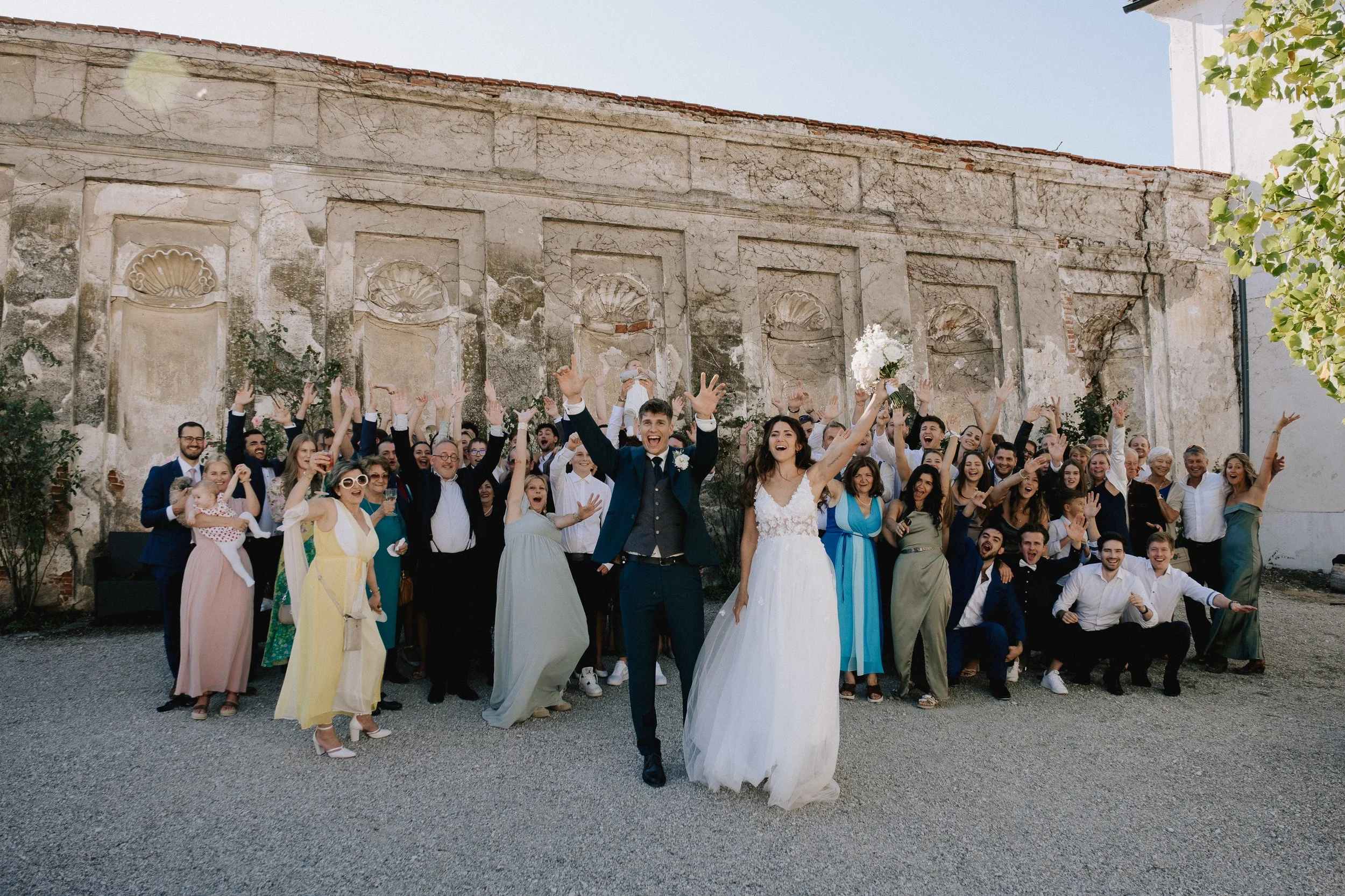 A large group of people, including a bride and groom, celebrating outdoors in front of a historic stone wall. The bride is holding a bouquet and smiling, while the groom is dressed in a suit. The guests are cheering and raising their hands in celebra