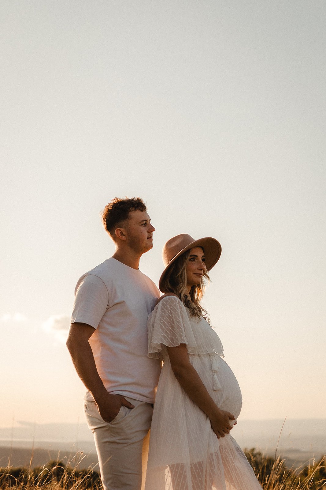 A couple, with the woman visibly pregnant, standing outdoors during sunset, facing to the right, with a clear sky and tall grass in the background.