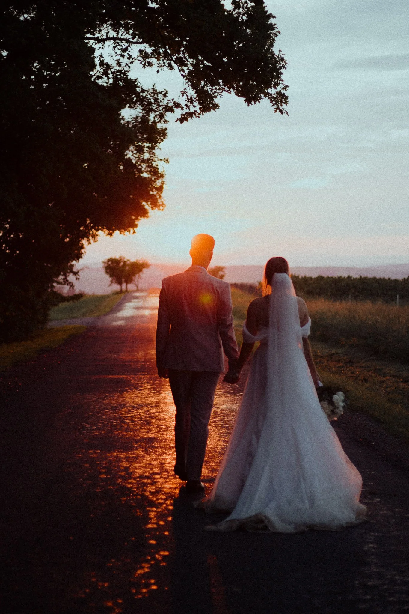 A bride and groom walking hand in hand on a rural road during sunset, with the bride wearing a white wedding gown and veil, and the groom in a suit.