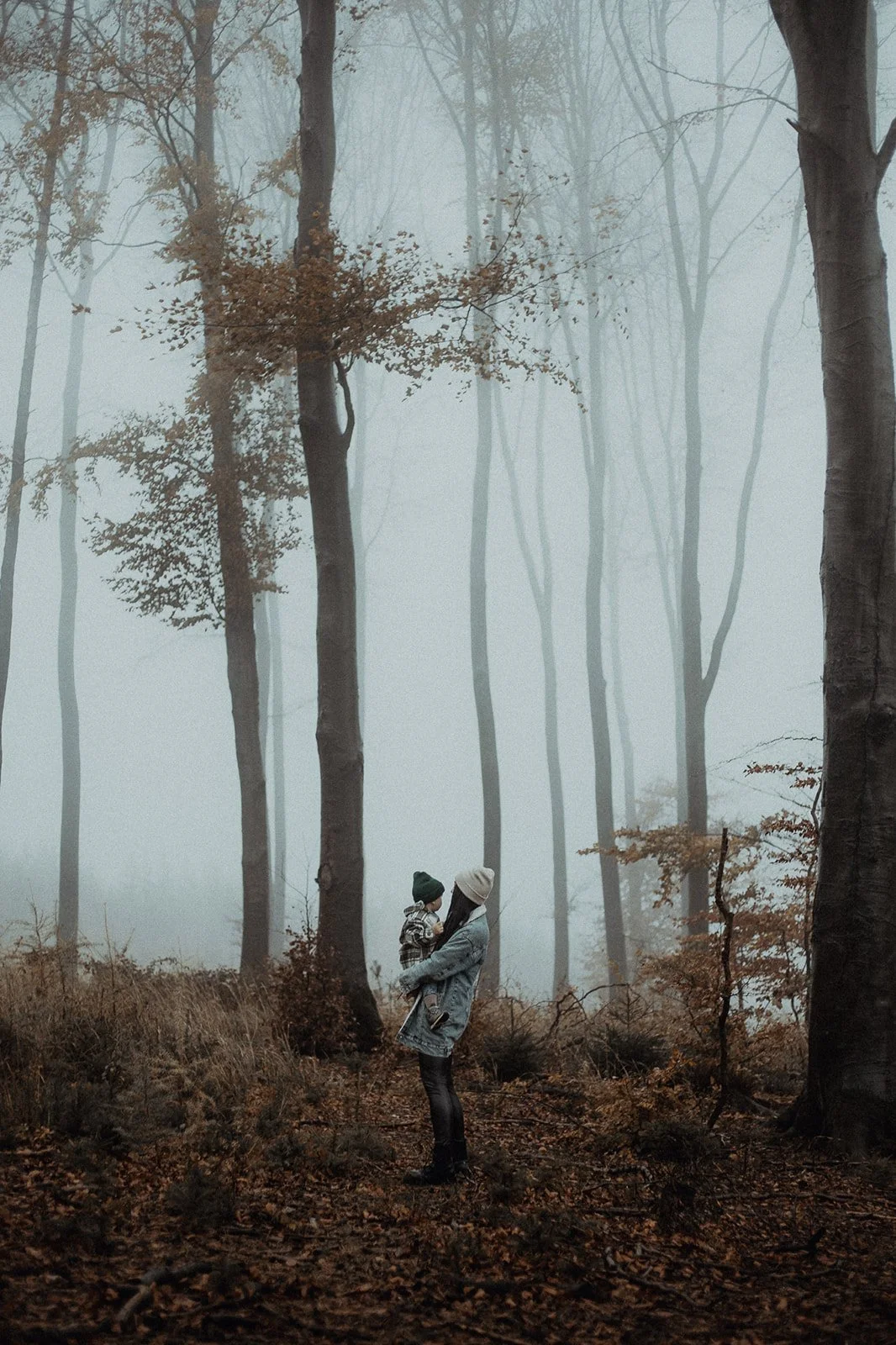 A woman holding a child in a foggy forest with tall trees and fallen leaves.