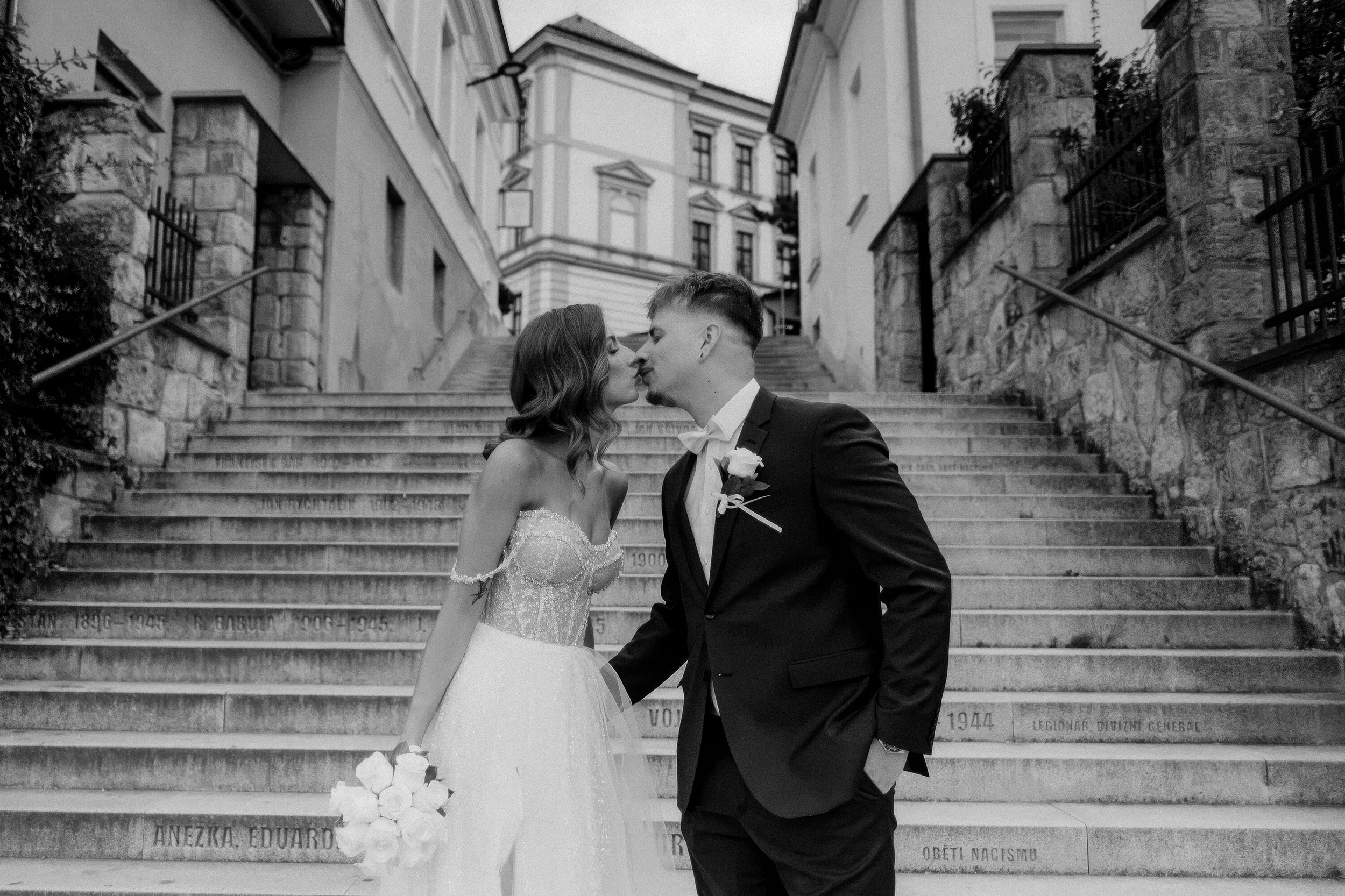 A black and white photo of a bride and groom sharing a kiss on a staircase outside a building, with the bride holding a bouquet of flowers.