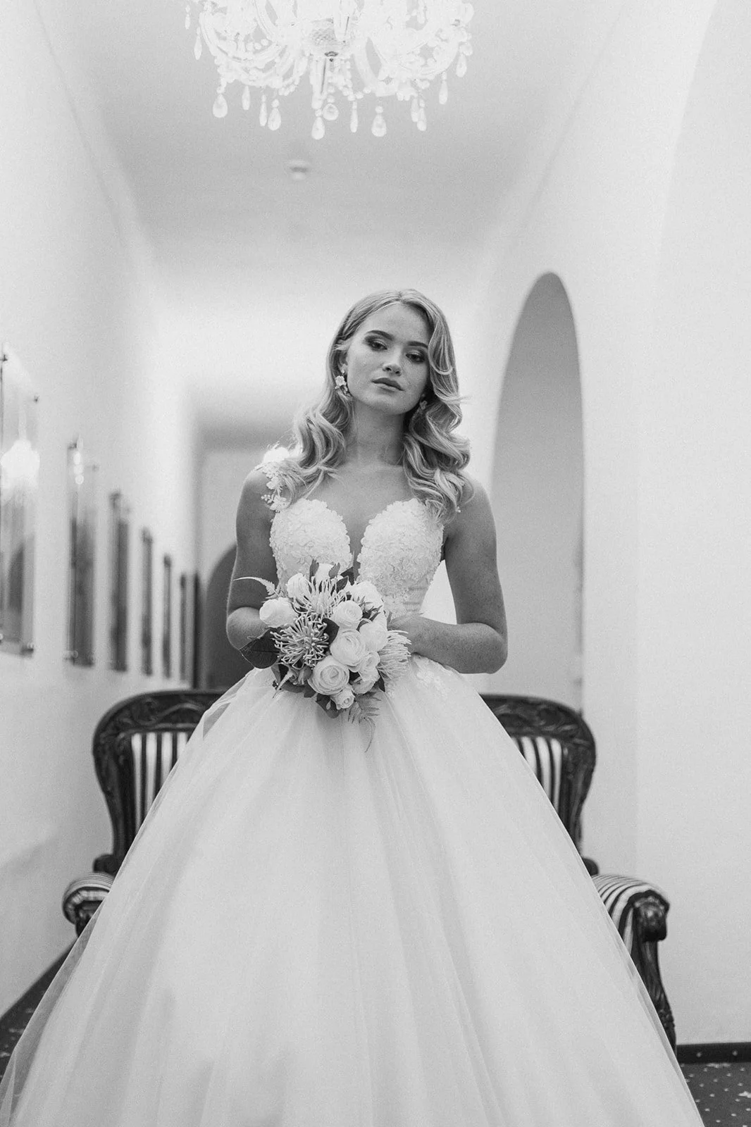 A bride in a wedding dress holding a bouquet, standing in an elegant hallway with chandeliers and framed pictures.