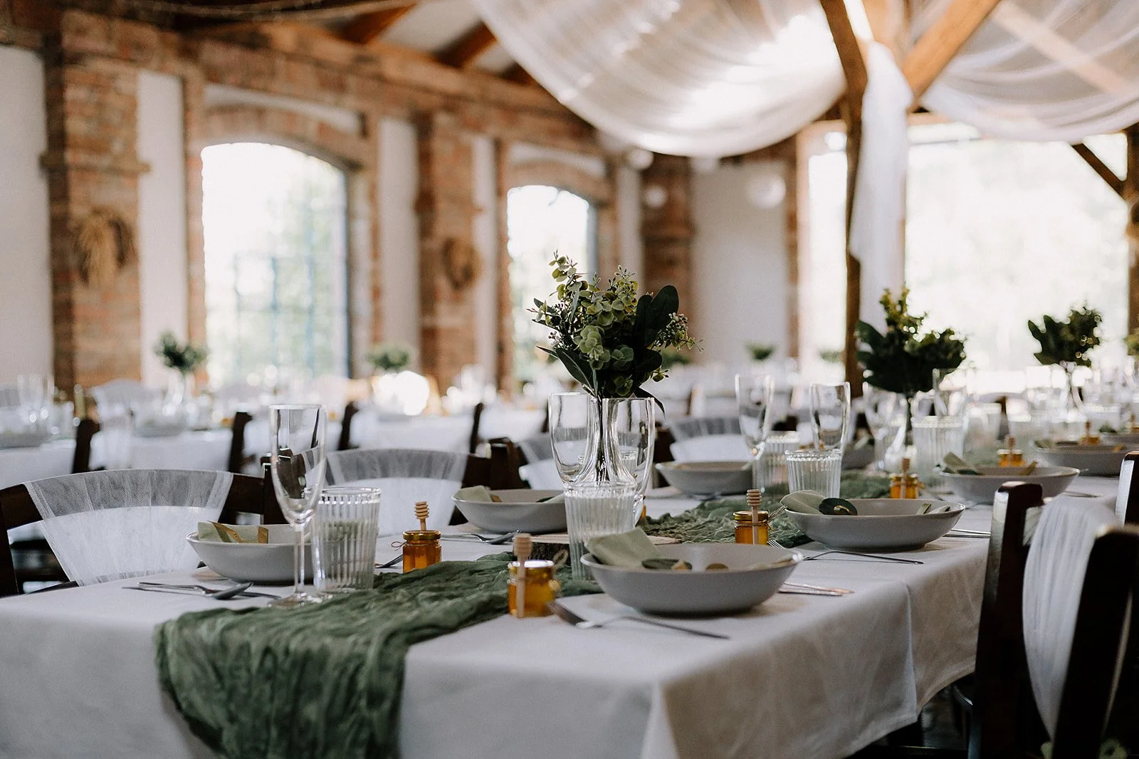 Empty wedding or banquet reception hall with decorated tables featuring floral centerpieces and tableware, bright natural lighting, and rustic brick walls.