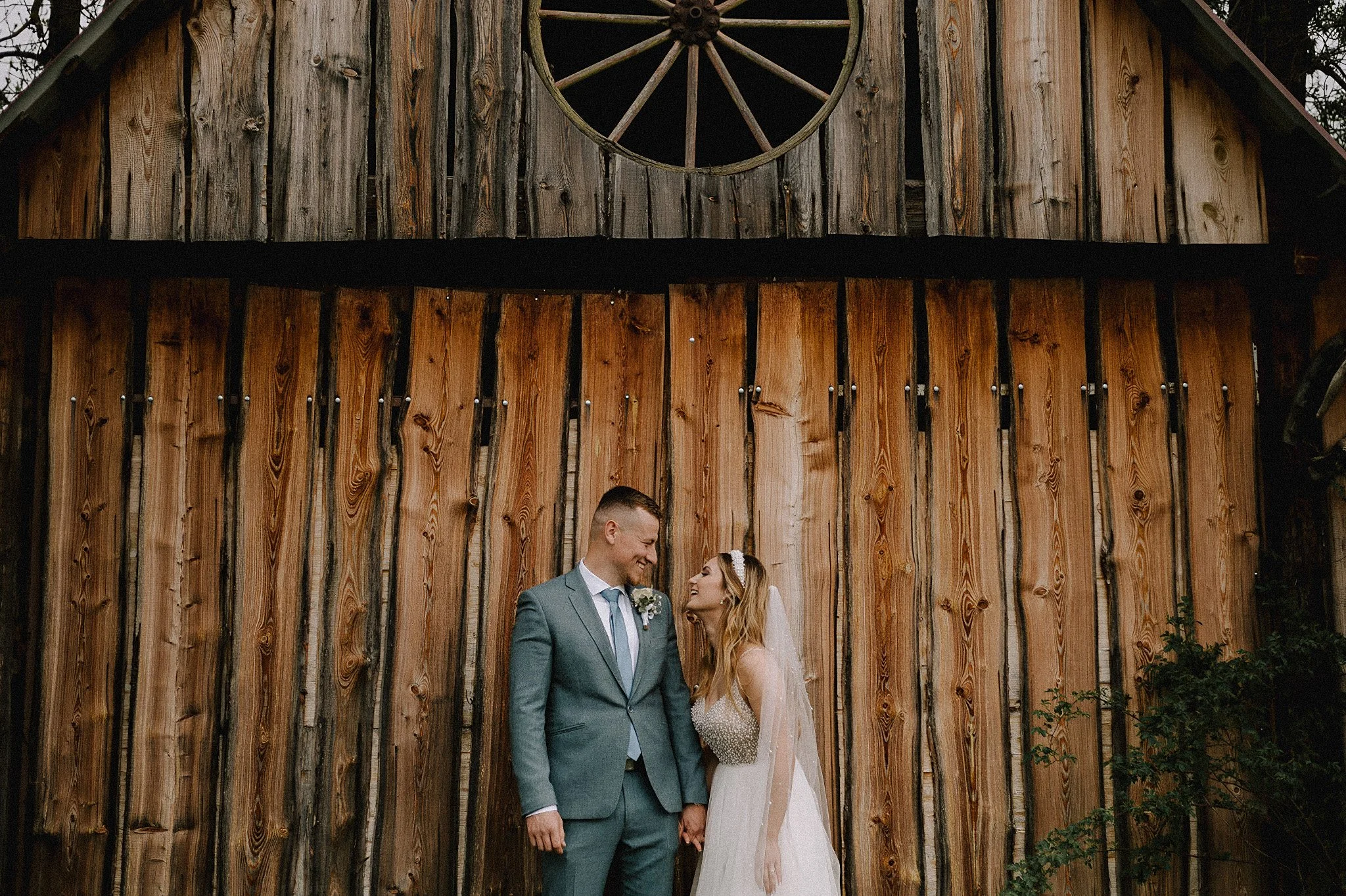 A bride and groom stand close together, holding hands, outside a rustic wooden barn with a decorative wheel above.