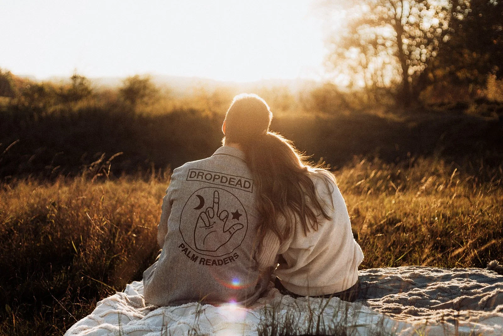 A couple sitting on a blanket in a grassy field during sunset, with their backs to the camera. The person on the left is wearing a jacket with a graphic and text on the back.