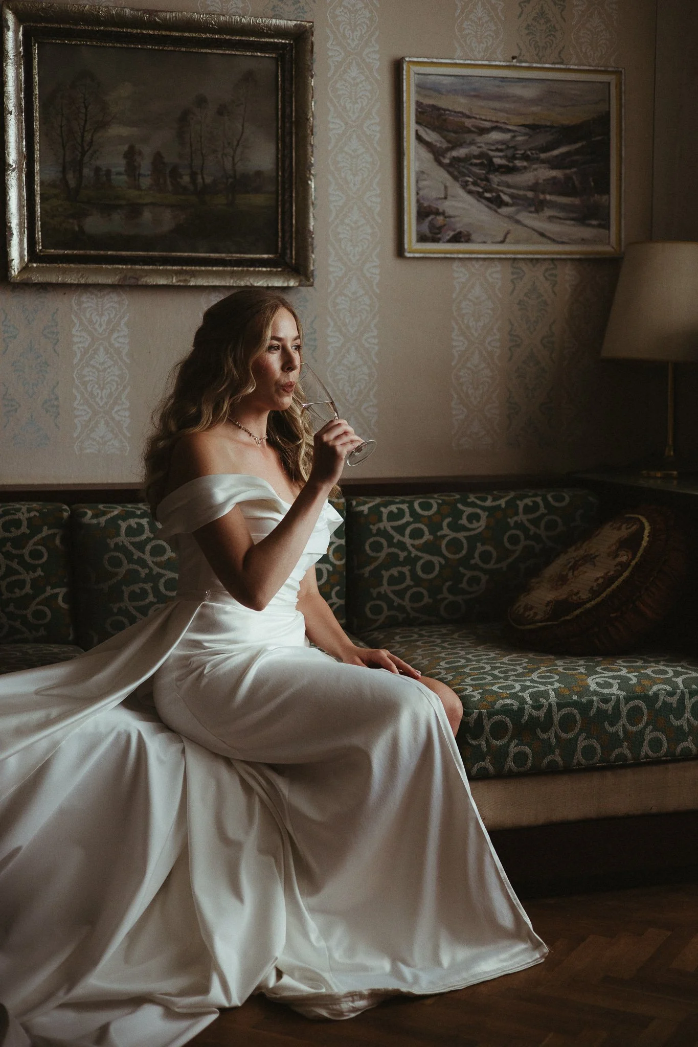 A woman in a white wedding dress sitting on a patterned couch, drinking from a wine glass in a dimly lit room with paintings on the wall.