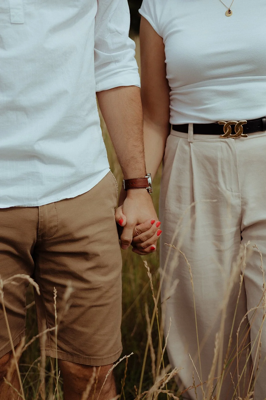 Close-up of a couple holding hands in a field of tall grass, with the woman wearing a white top and beige pants, and the man wearing a white shirt and light brown shorts.