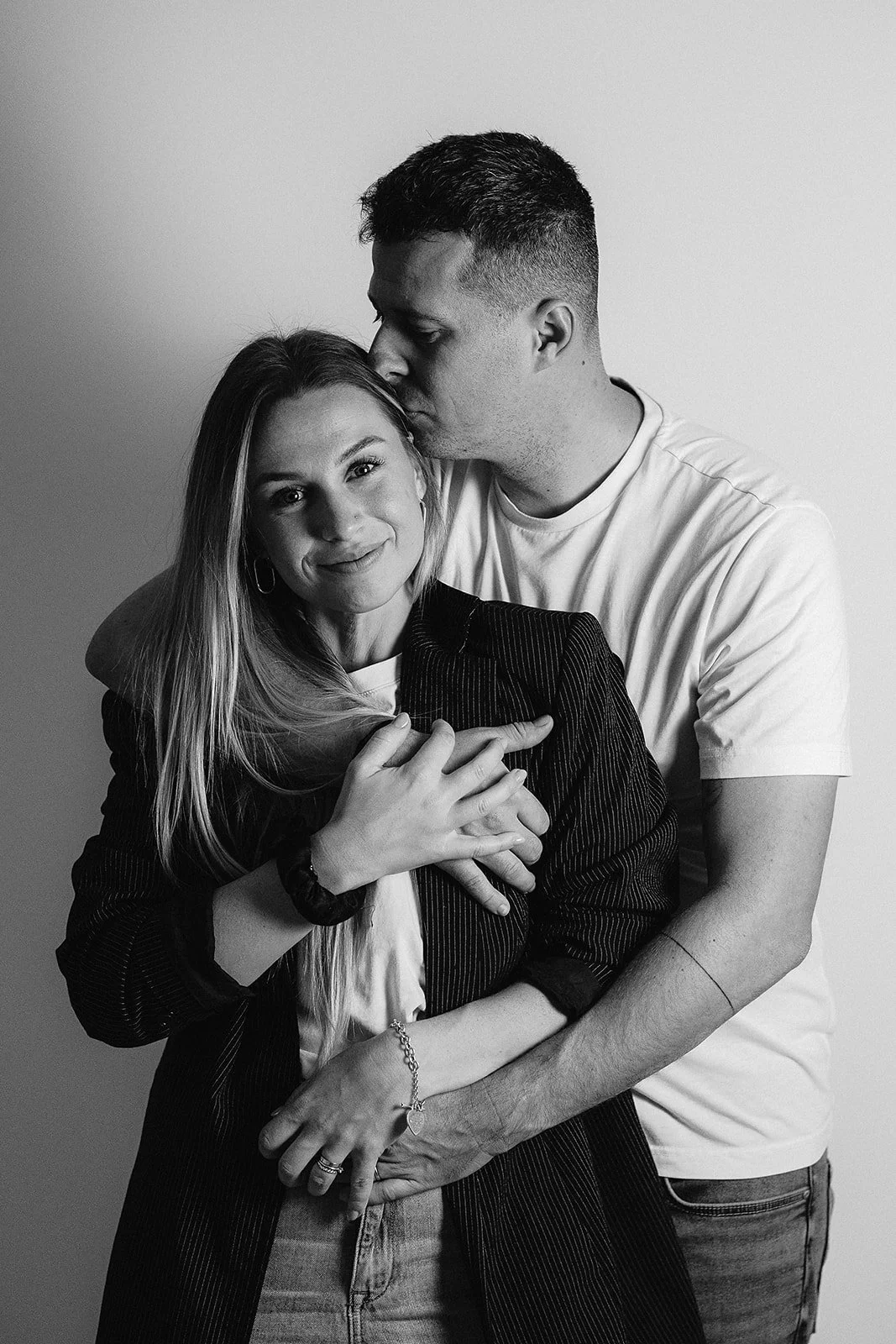 A black and white photo of a couple embracing. The man is kissing the woman's forehead while she smiles gently, with her hand on his chest and her arm around his waist.