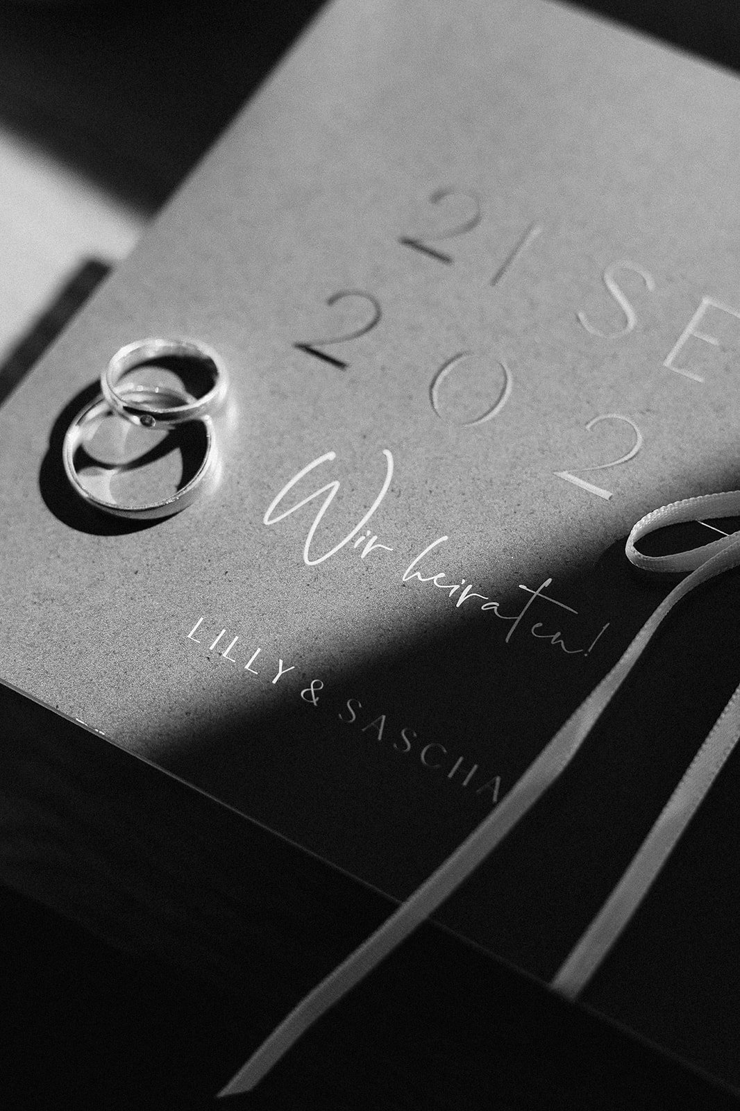 A black and white photograph of an invitation card for a wedding, with two wedding rings placed on top. The text on the card includes the words "save the date," the date "22.09.23," and the phrase "Wir heiraten!" along with the names Lilly and Saskia