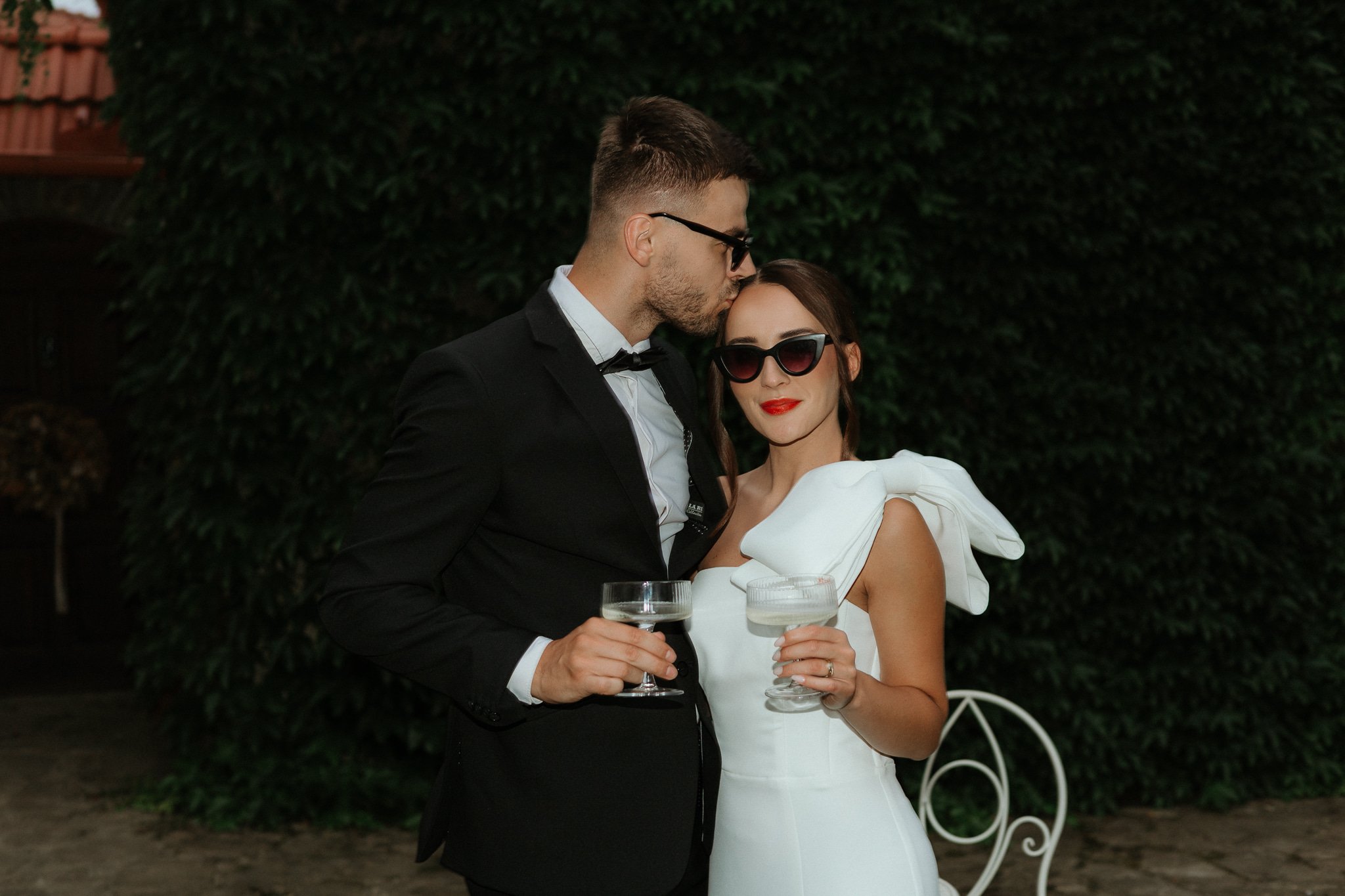 A stylish couple dressed in formal attire, the man in a black tuxedo with a bow tie and the woman in a white dress with prominent shoulder bow details, holding cocktails and standing close together in front of greenery at an outdoor event.