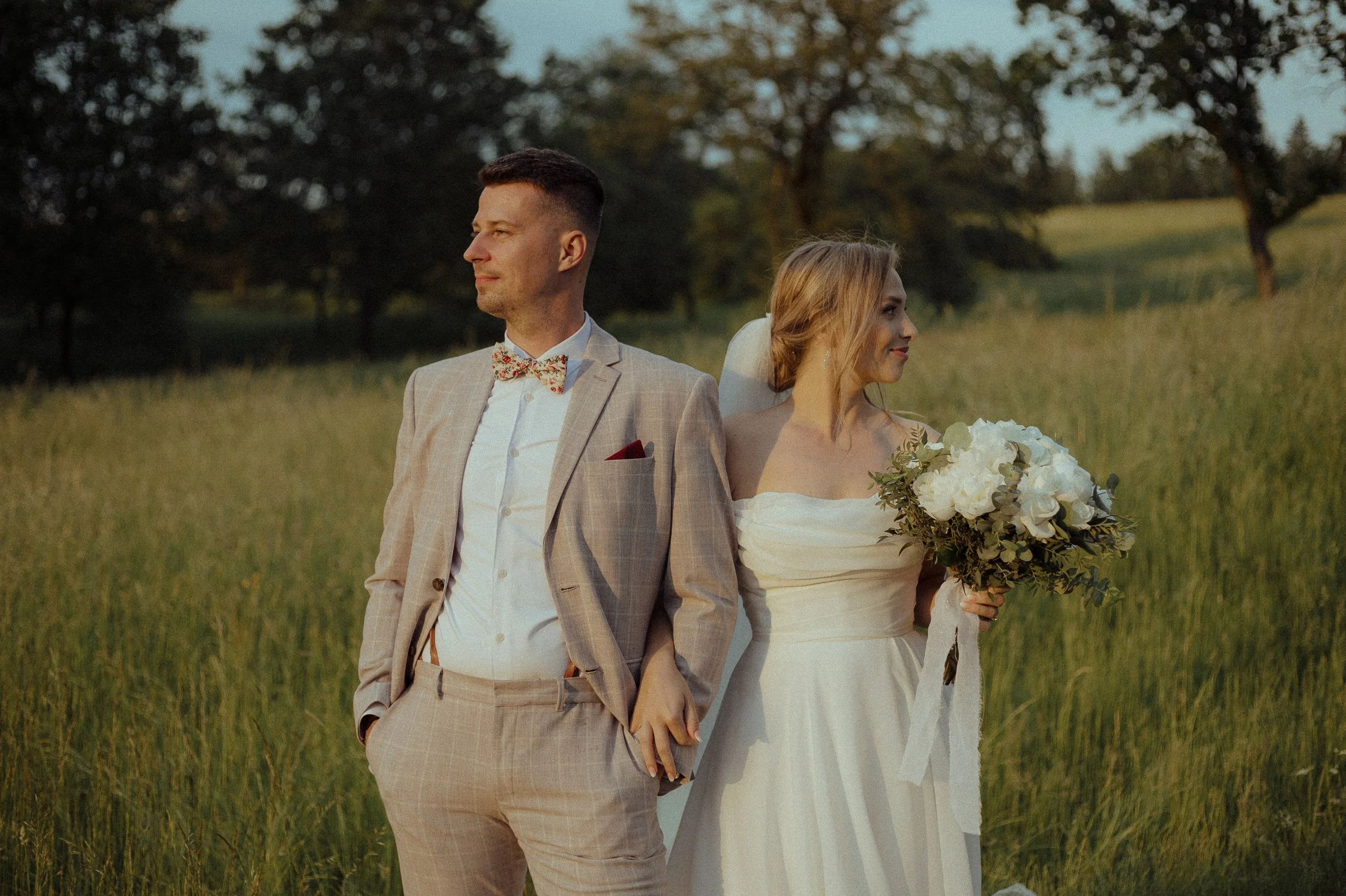 A bride and groom holding hands in a grassy field at sunset, the groom in a light beige plaid suit with a floral bowtie, and the bride in a strapless white wedding gown holding a bouquet of white roses, with trees in the background.