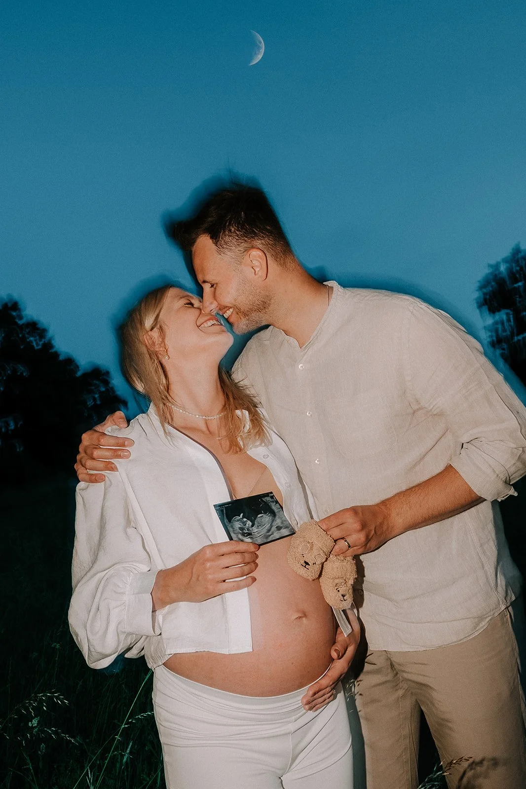 Pregnant woman and man smiling and touching foreheads outdoors at dusk, holding sonogram picture, teddy bear, and a sonogram ultrasound scan, with moon in the sky.