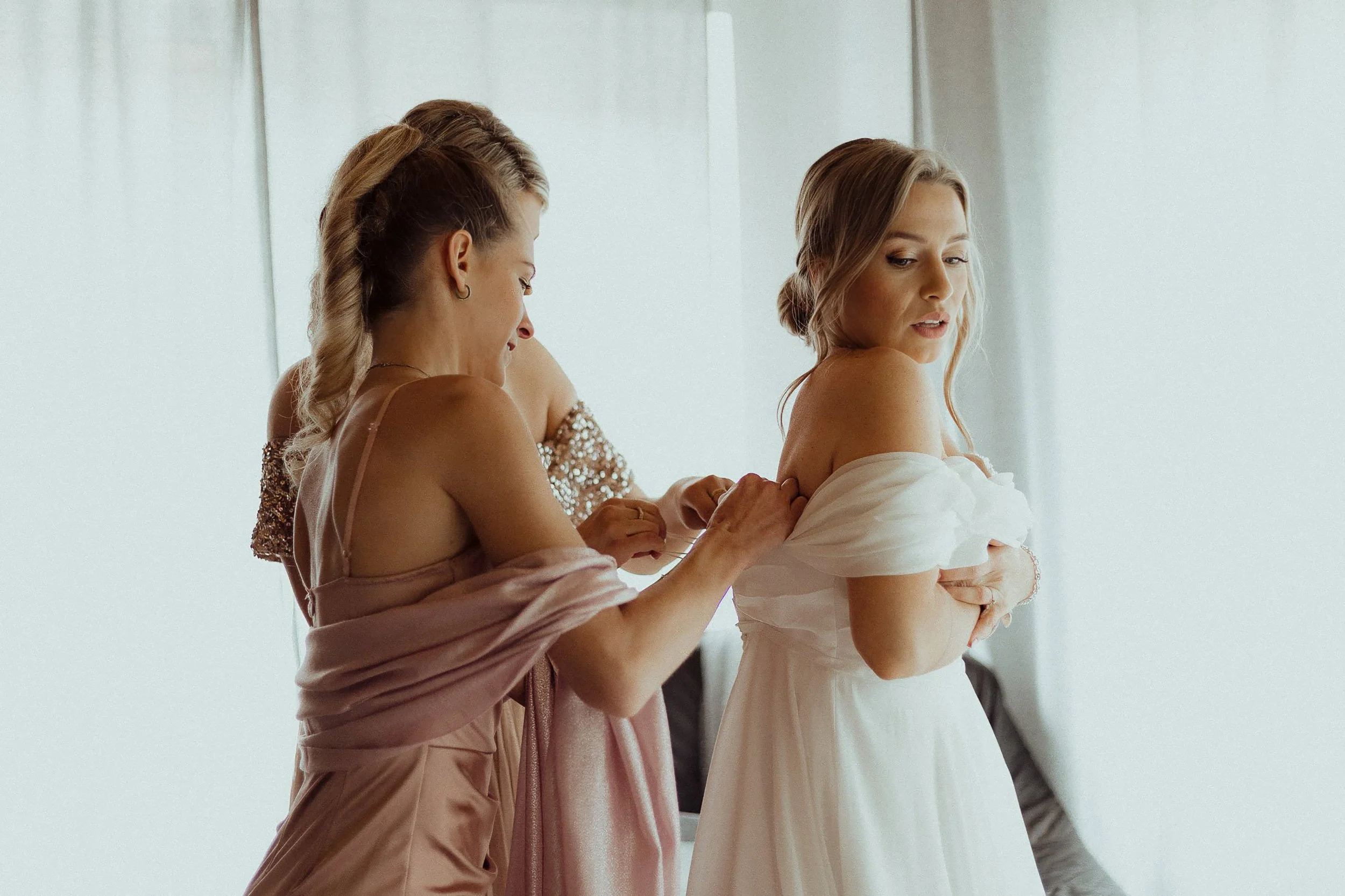 Two women helping a bride get dressed in a white gown, with one woman adjusting the dress's shoulder straps. The bride looks to the side, and the room is bright and airy.