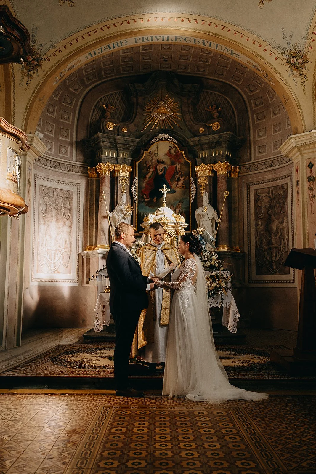 A wedding ceremony taking place in a church with a couple standing in front of an altar, holding hands and exchanging vows with a priest officiating. The bride is wearing a white lace wedding gown and the groom a dark suit. The church interior is orn