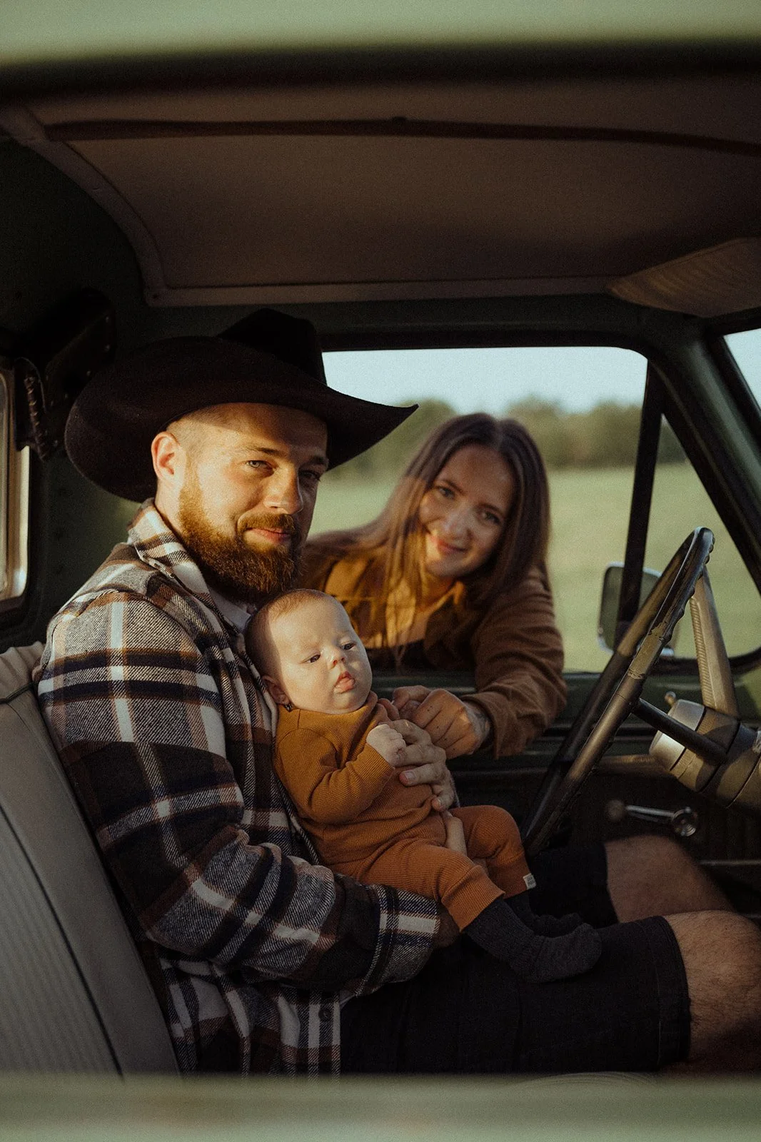 A family of three sitting inside a vehicle, with the father holding a baby, the mother in the background smiling, all dressed in casual clothing.