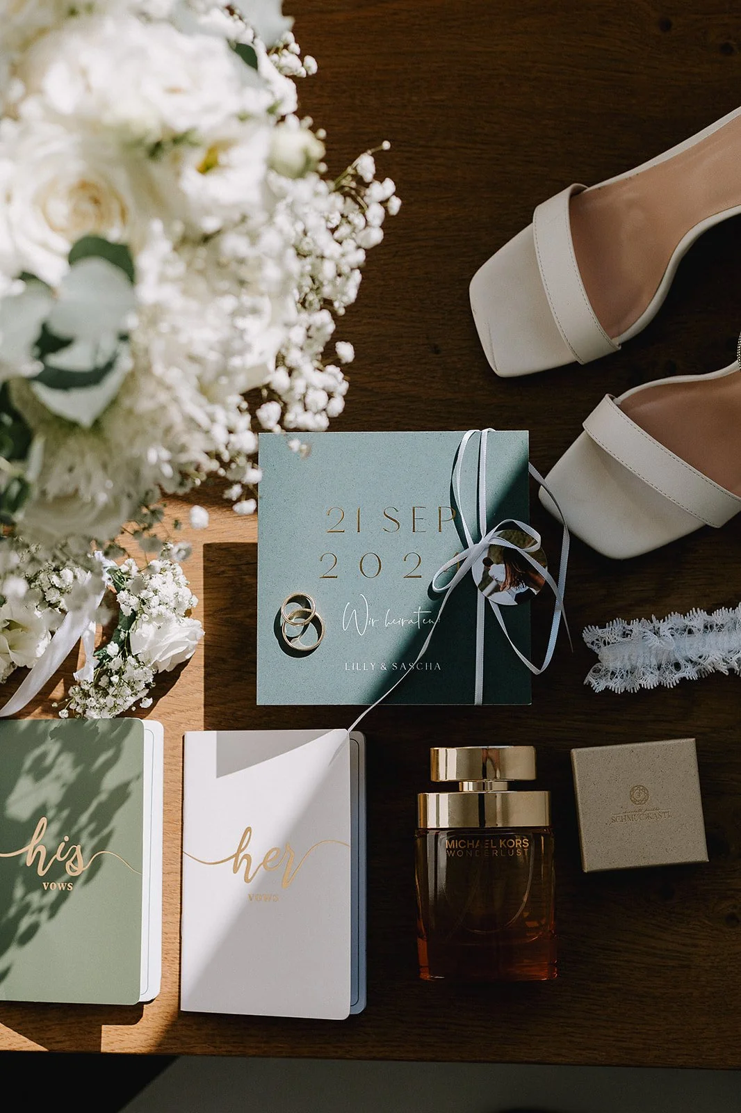 Wedding invitation suite with rings and bouquet of white flowers, white high-heeled shoes, a box of perfume, and vows cards on a wooden surface.