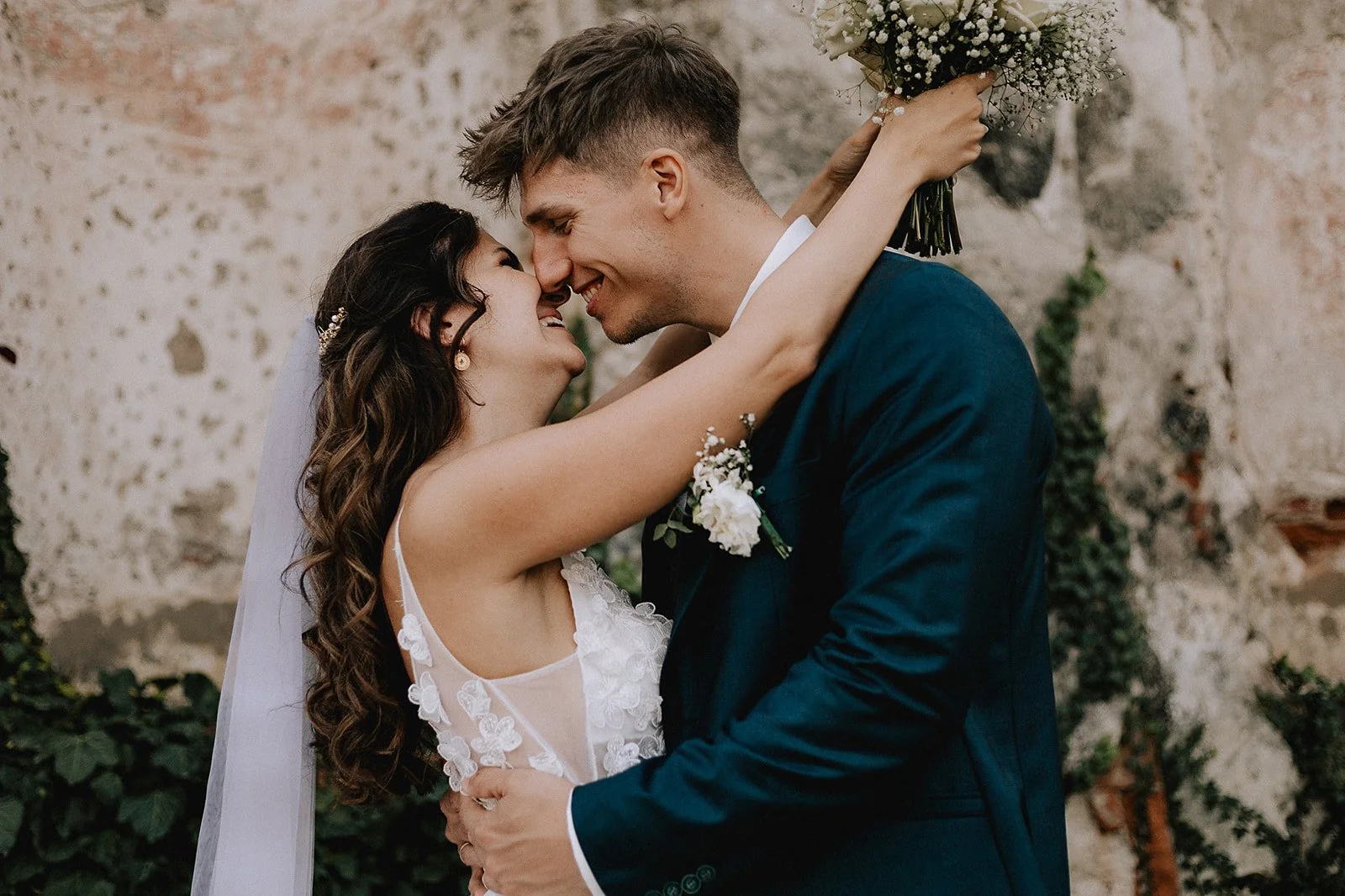 A bride and groom close together, touching foreheads, smiling with noses almost touching, in wedding attire with a rustic background.