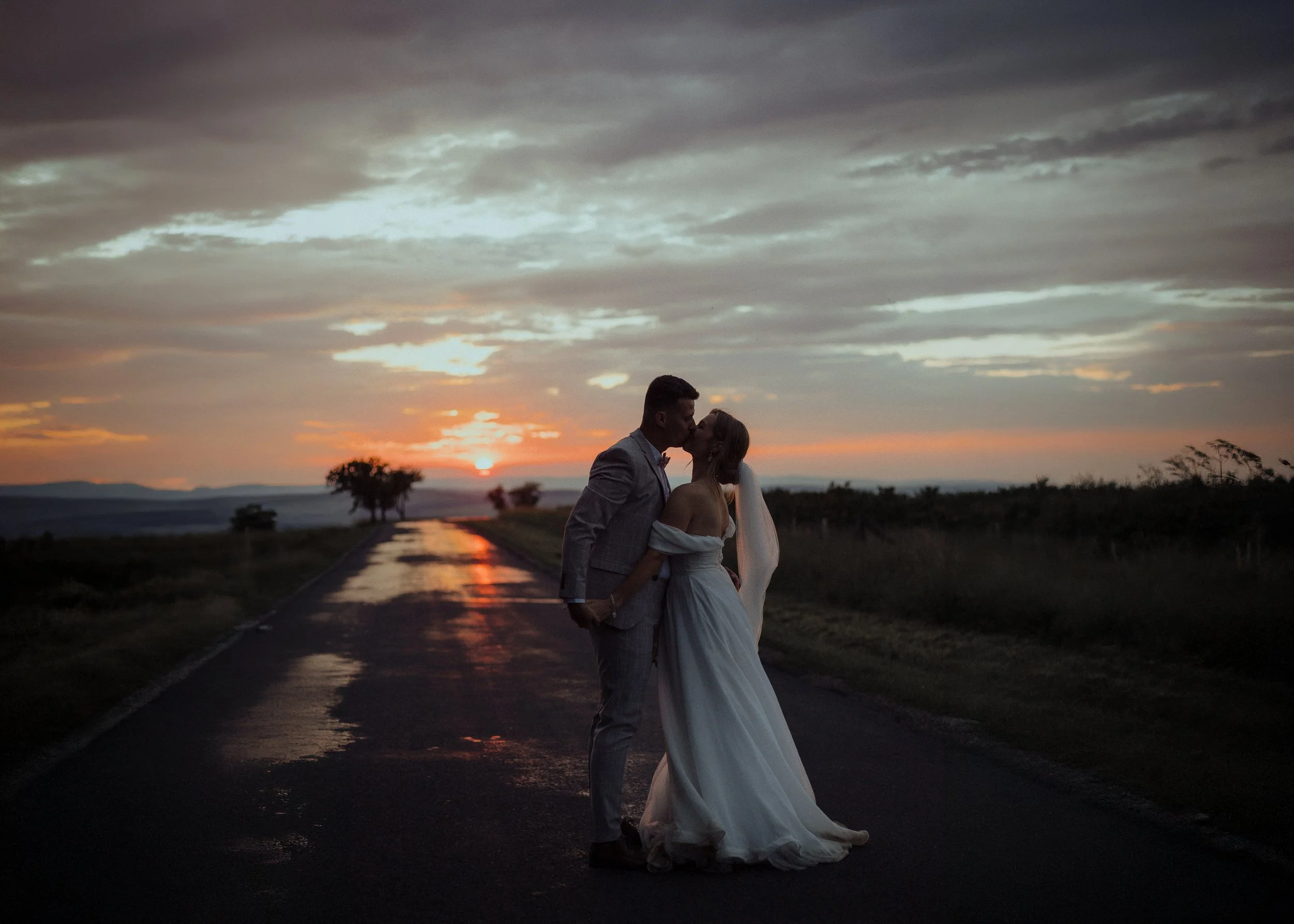 A bride and groom kissing on a rural road during sunset, with trees in the background.