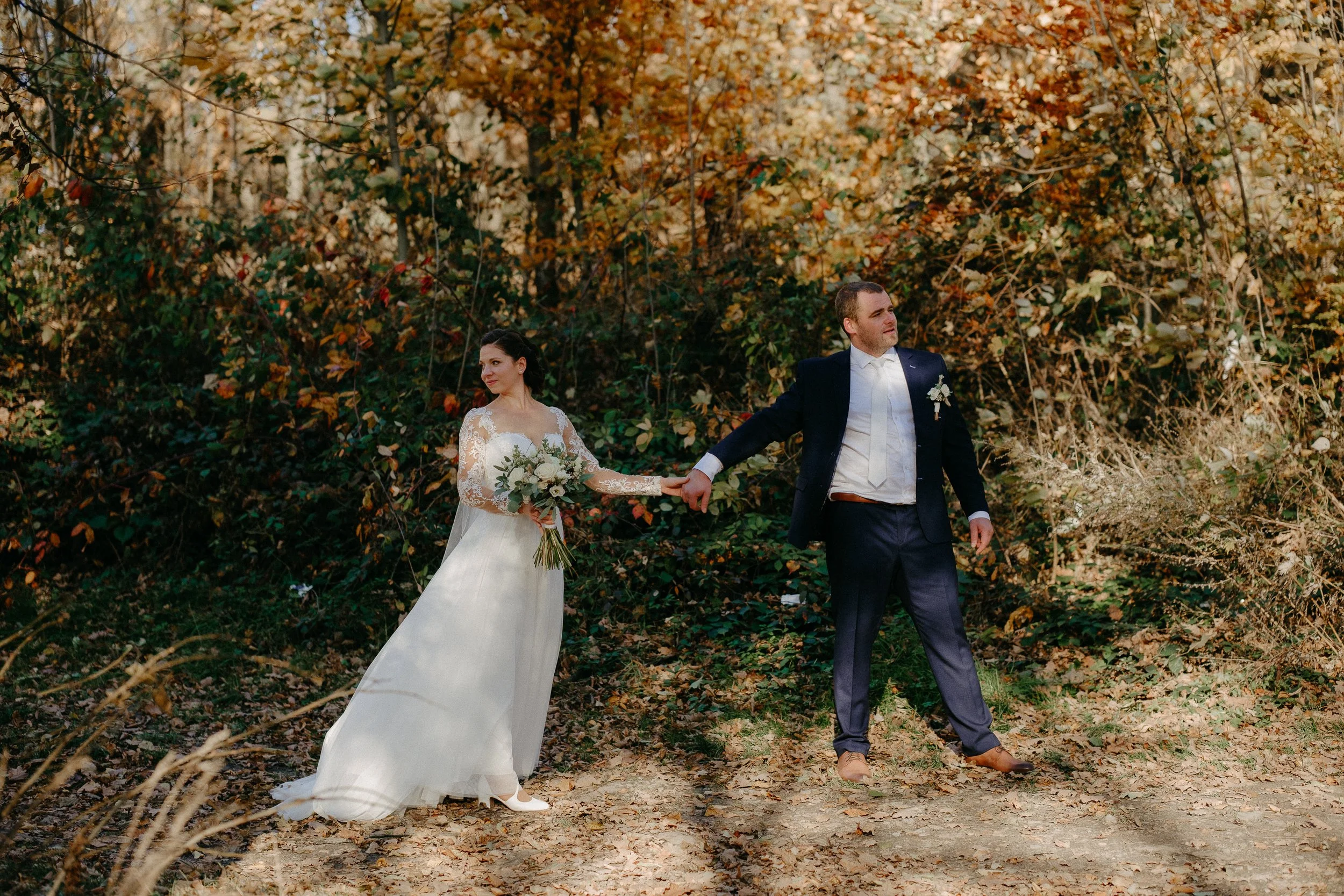 Couple outdoors in a forest, bride holding bouquet and wearing a white wedding dress with lace sleeves, groom in a dark suit, holding hands with bride, autumn leaves in background.