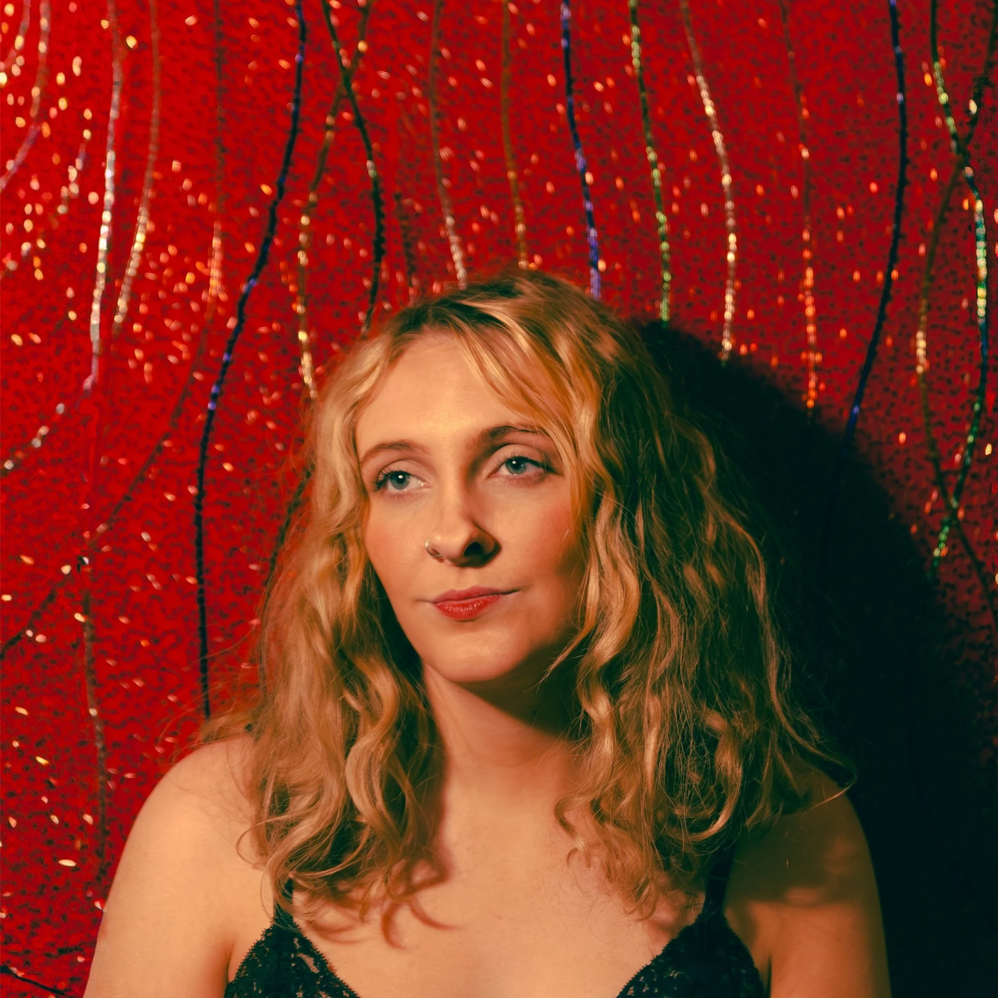 A woman with long curly blonde hair wearing a black top against a red sequin curtain background.